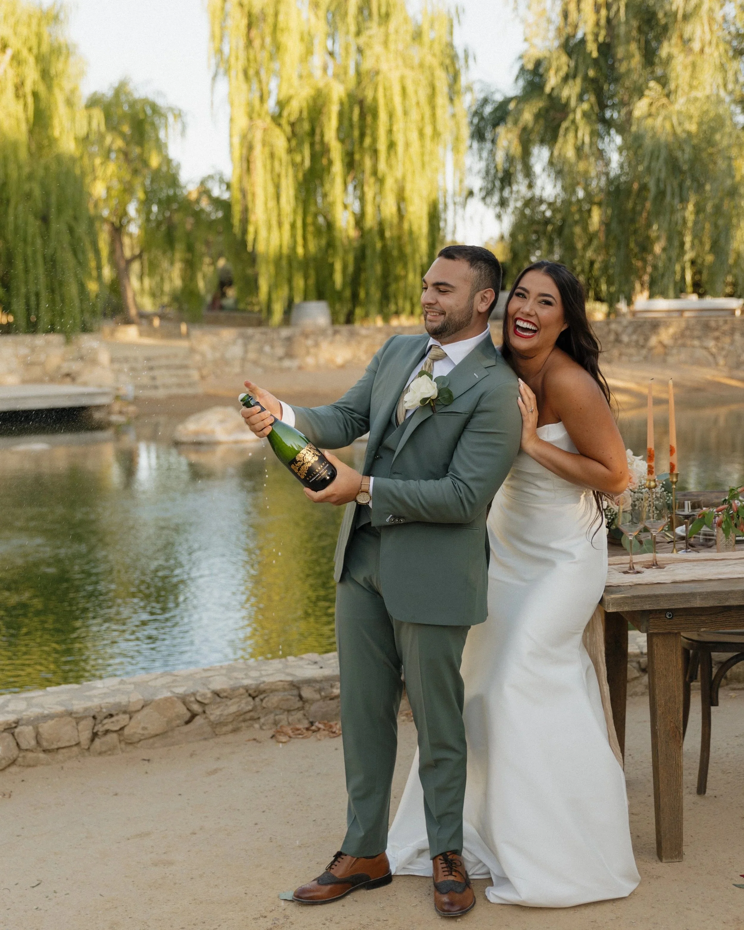 A happy couple in wedding attire enjoying a outdoor moment by a pond. The groom is holding a bottle of champagne while the bride is smiling and embracing him from behind. The background features a pond, trees, and outdoor tables, suggesting a wedding celebration.