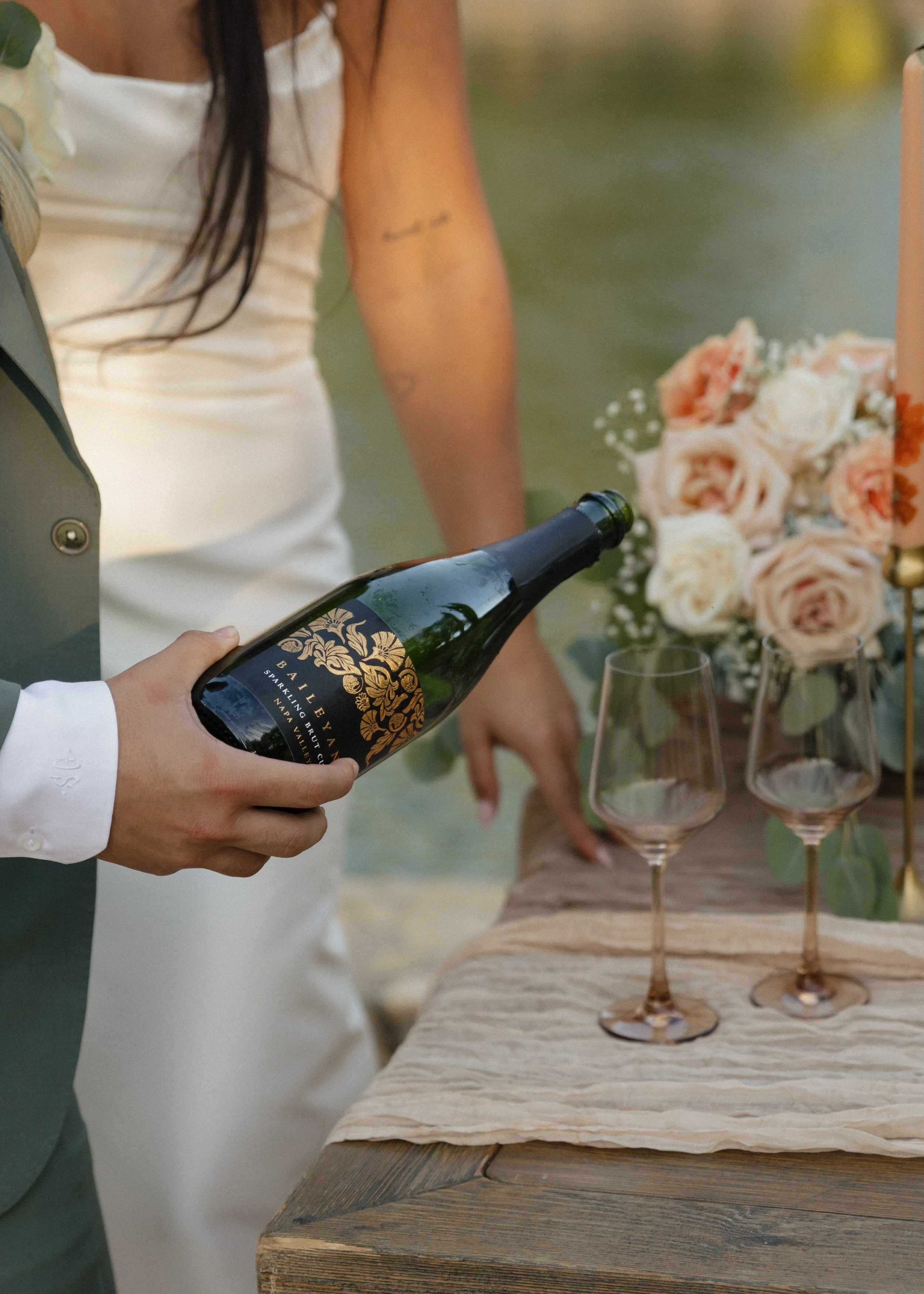 Person pouring champagne into glasses at a wedding or celebration setting with flowers on a wooden table.