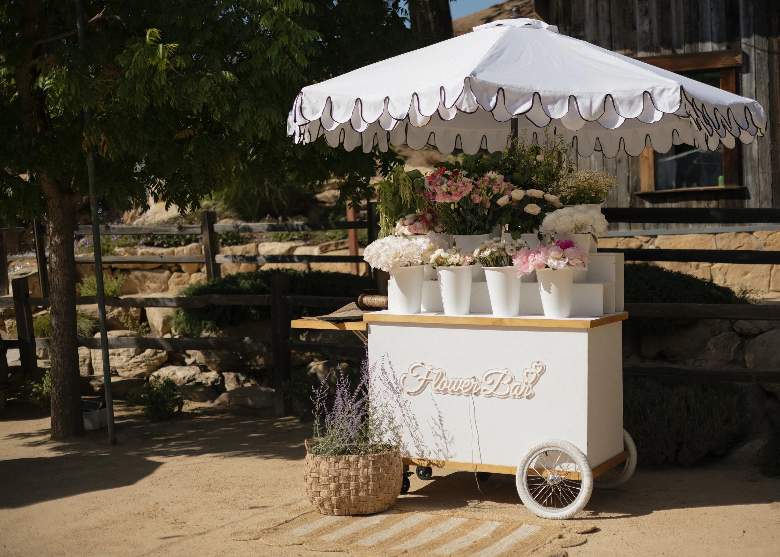 A flower cart with a white umbrella, displaying various bouquets of pink, white, and purple flowers in white vases, with a wicker basket of lavender in front, against a rustic wooden building and trees.
