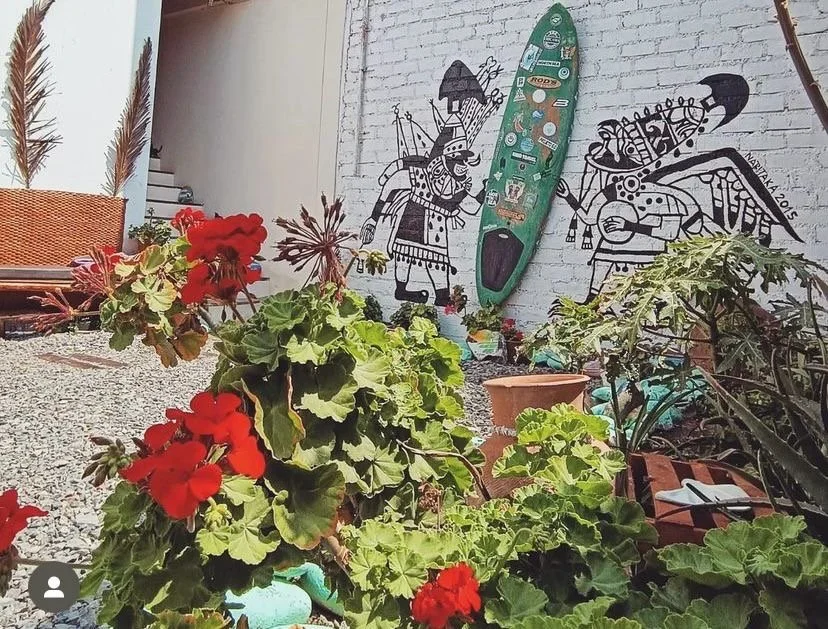 Jardín con plantas verdes y flores rojas, mural en la pared con figuras indígenas y una pala decorativa