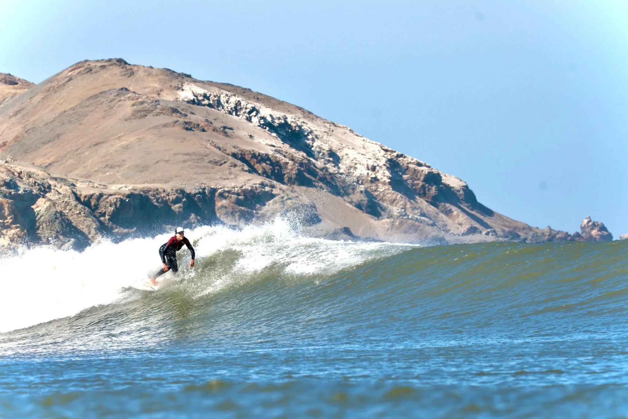 Una persona surfea en una ola en el mar con una montaña en el fondo.