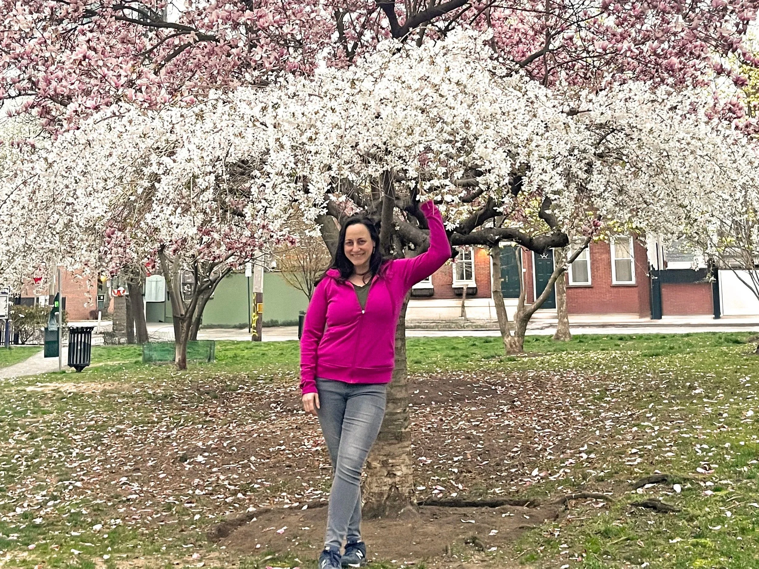 Woman with dark hair in pink top in front of a tree with flowers.