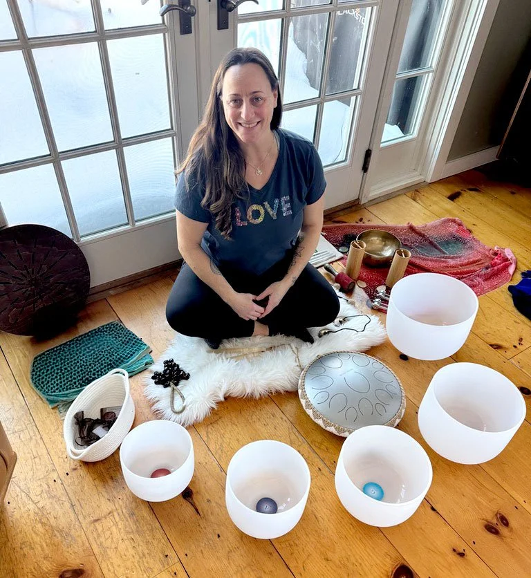 Image of a woman smiling sitting on a wood floor with singing bowls and other instruments around her.