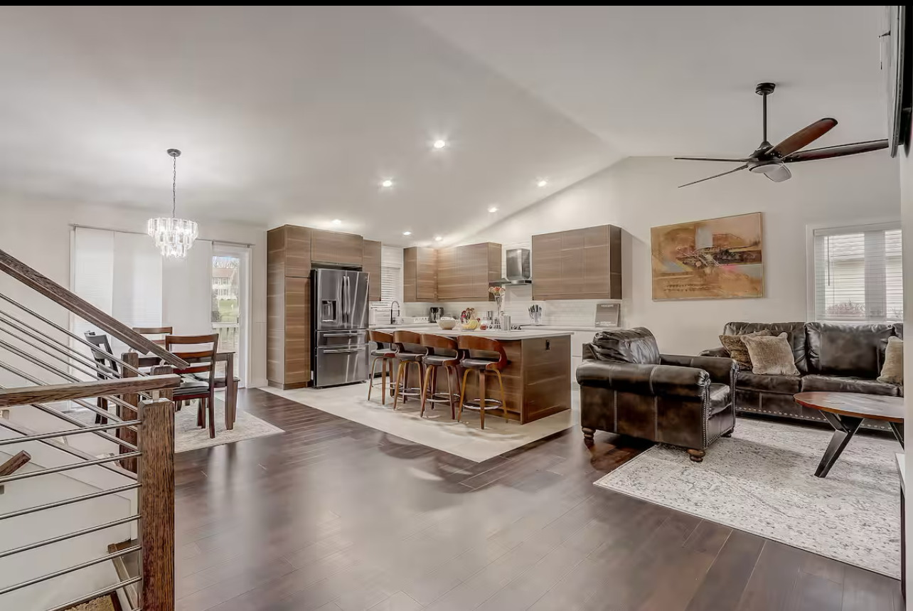Open-concept living room and kitchen with dark hardwood floors, a black leather sectional sofa, a wooden coffee table, and a kitchen island with barstools.