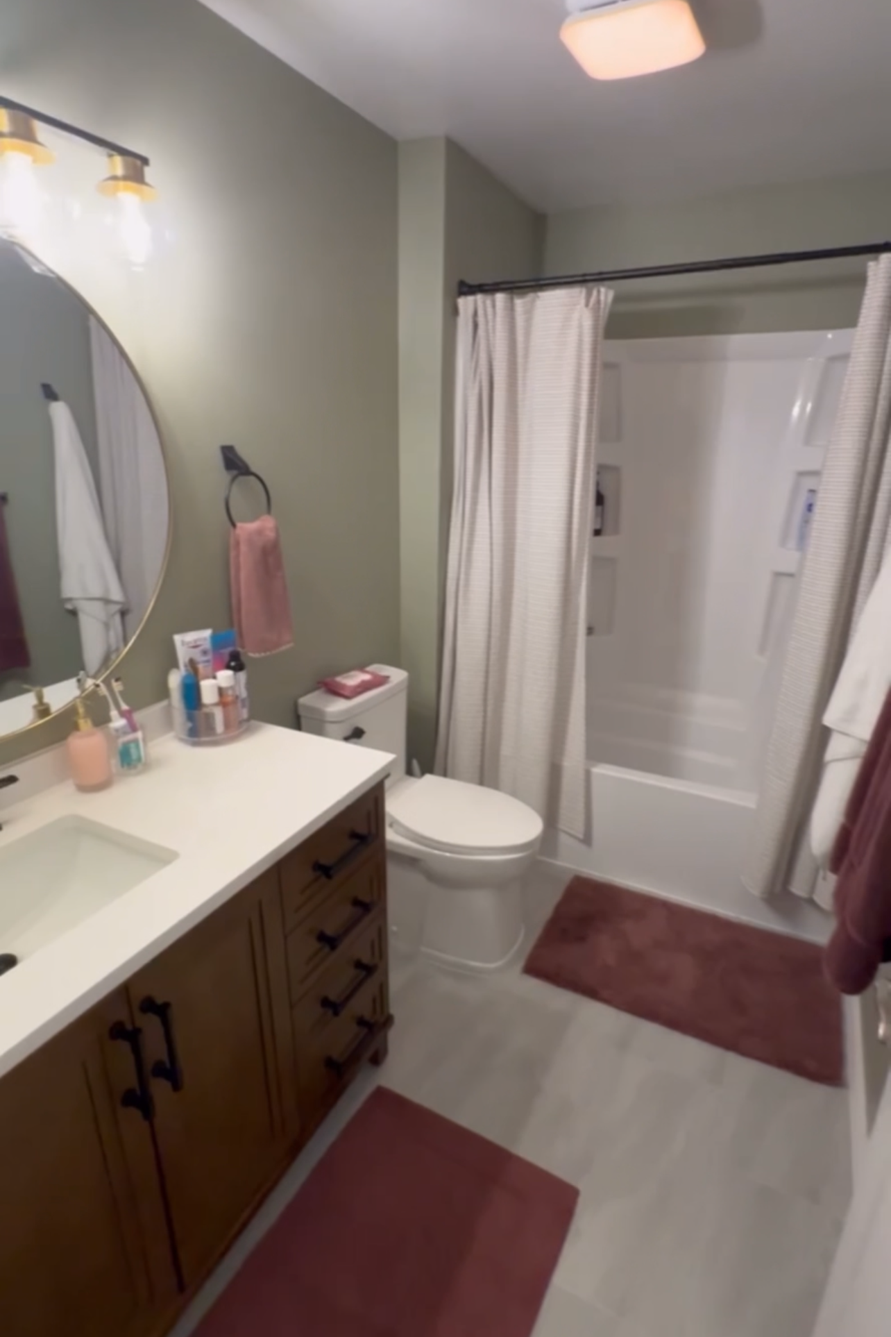 View of a small bathroom with a wooden vanity, a round mirror, a toilet, a bathtub with a shower curtain, and various toiletries on the countertop. There are pink towels hanging and on the floor.