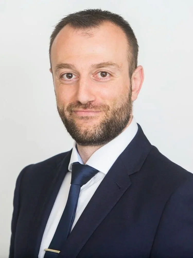 A professional headshot of a man in a dark suit, white shirt, and dark tie, with a light background.