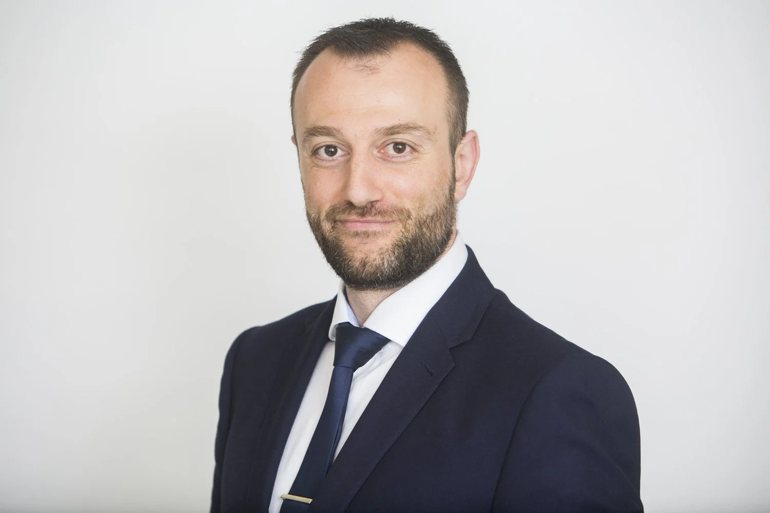 A man with short brown hair and a beard, dressed in a dark suit and tie, smiling at the camera against a plain white background.