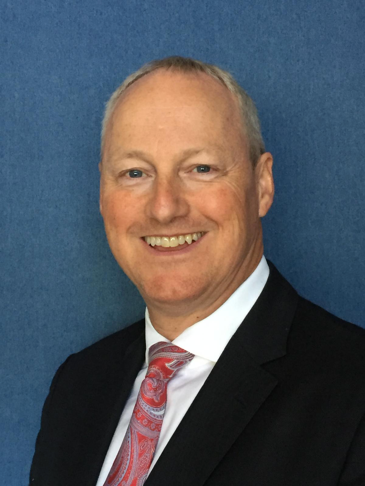 Photo of James A. Edens, P.Eng, Founder & Chief Executive Officer, Inceptio Oil & Gas Limited. A smiling man in a black suit, white shirt, and a red patterned tie, standing against a blue background.