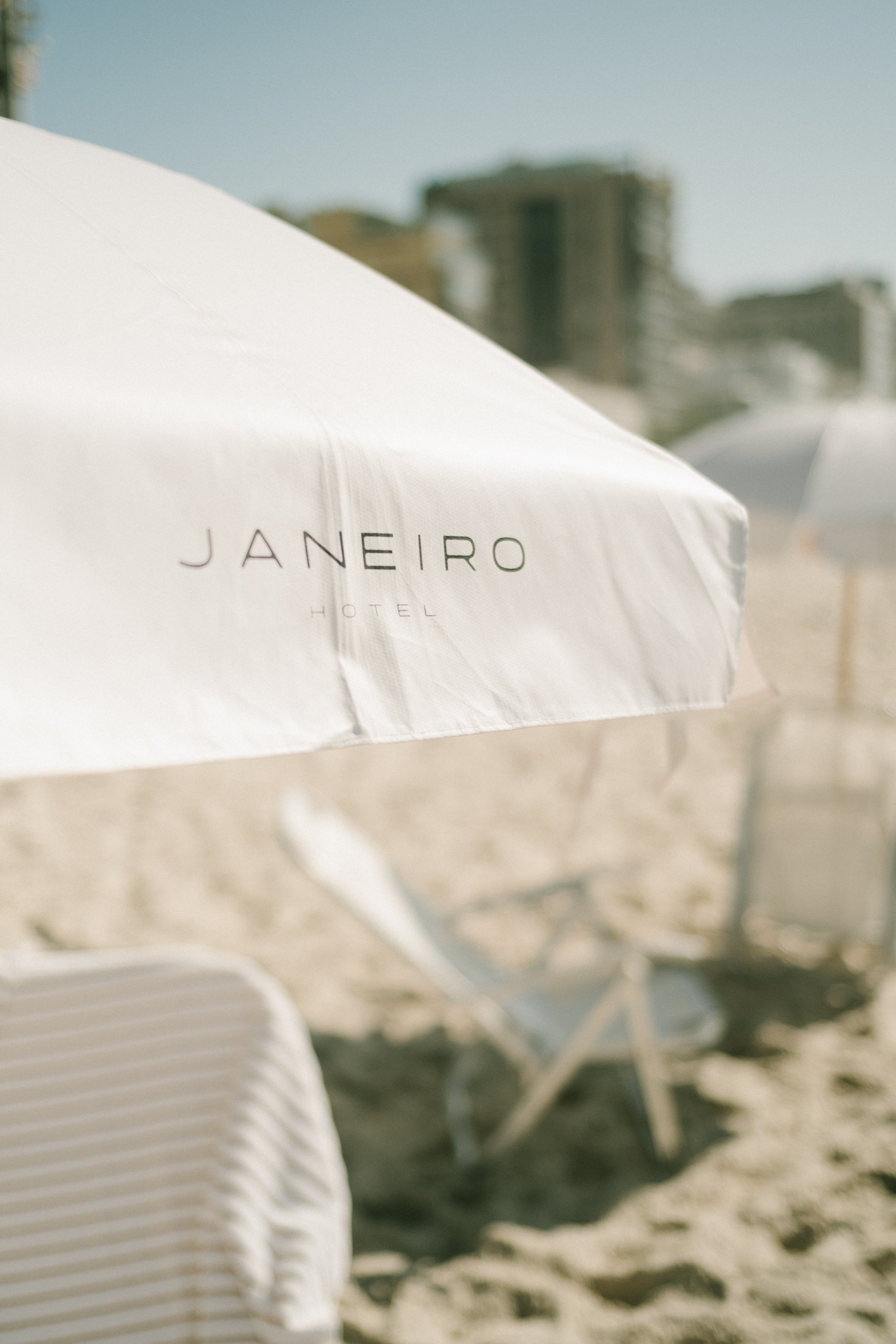 Uma sombra de guarda-sol branco na areia de uma praia, com fundo de edifícios altos e céu claro.
