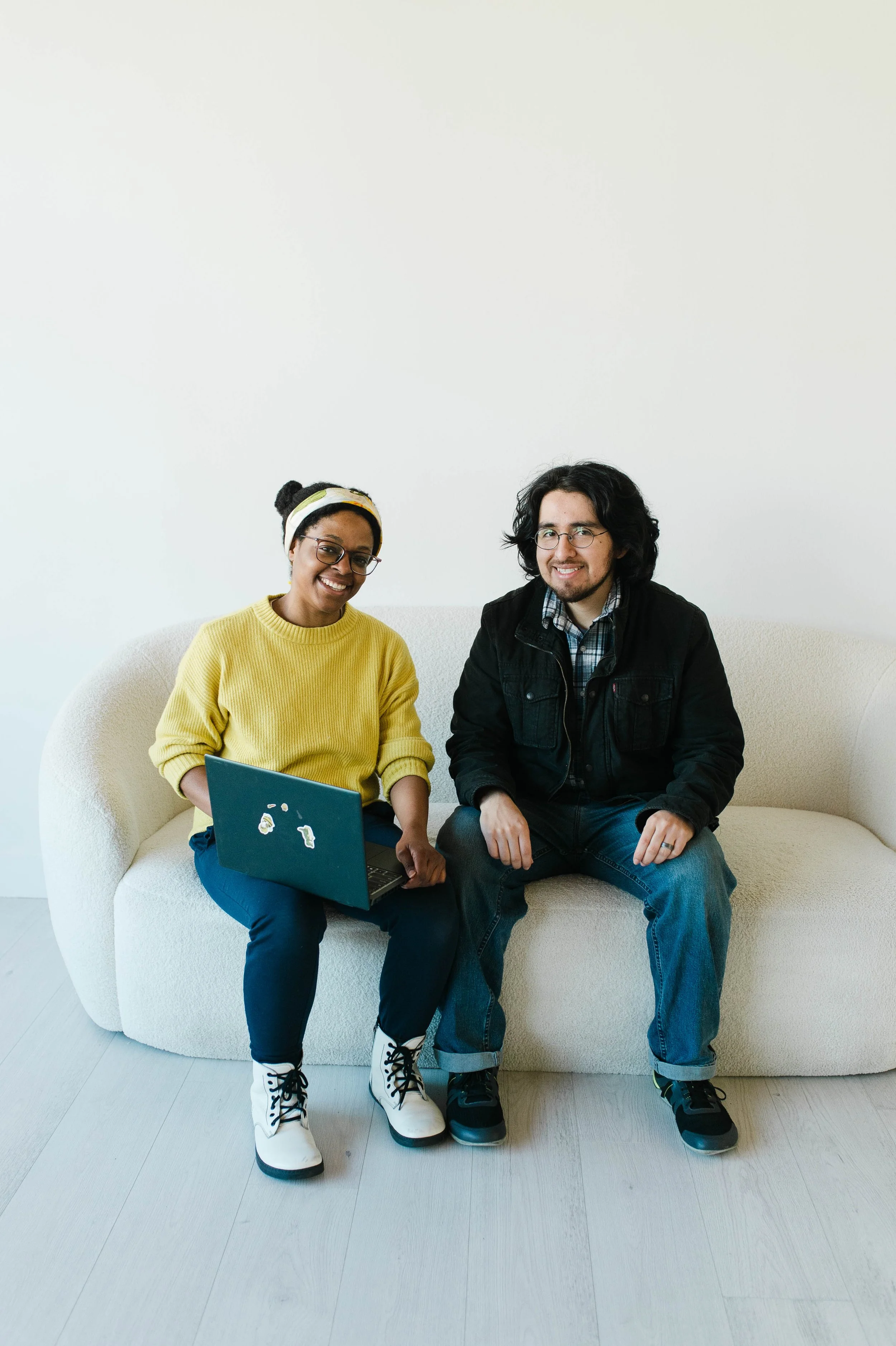 Aneisha, black woman wearing a yellow sweater, shows laptop screen to Aaron, man with shoulder length black hair and black jacket. They both smile.