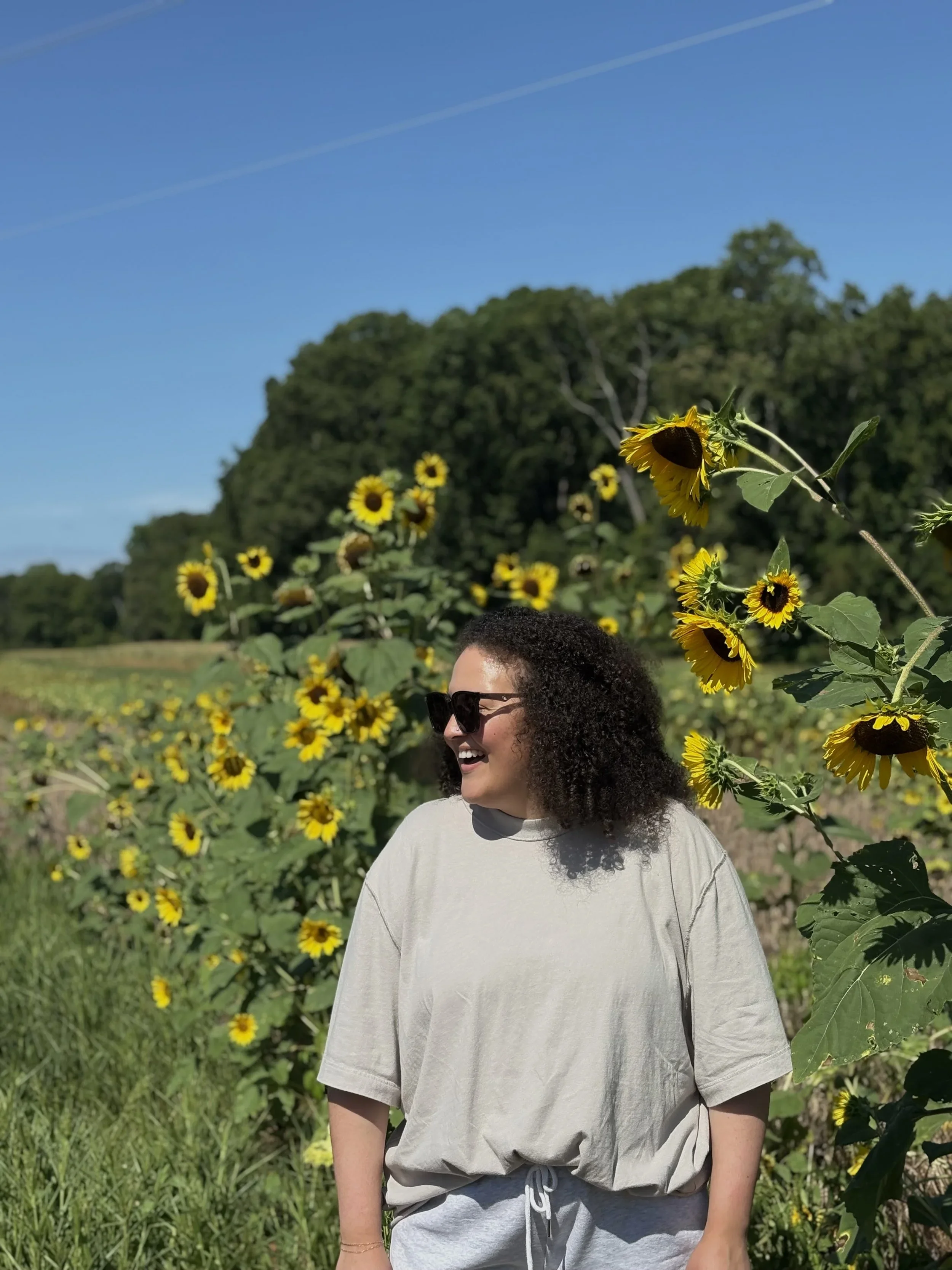 Andrea stands in front of sunflowers outside, looking to the side and smiling.