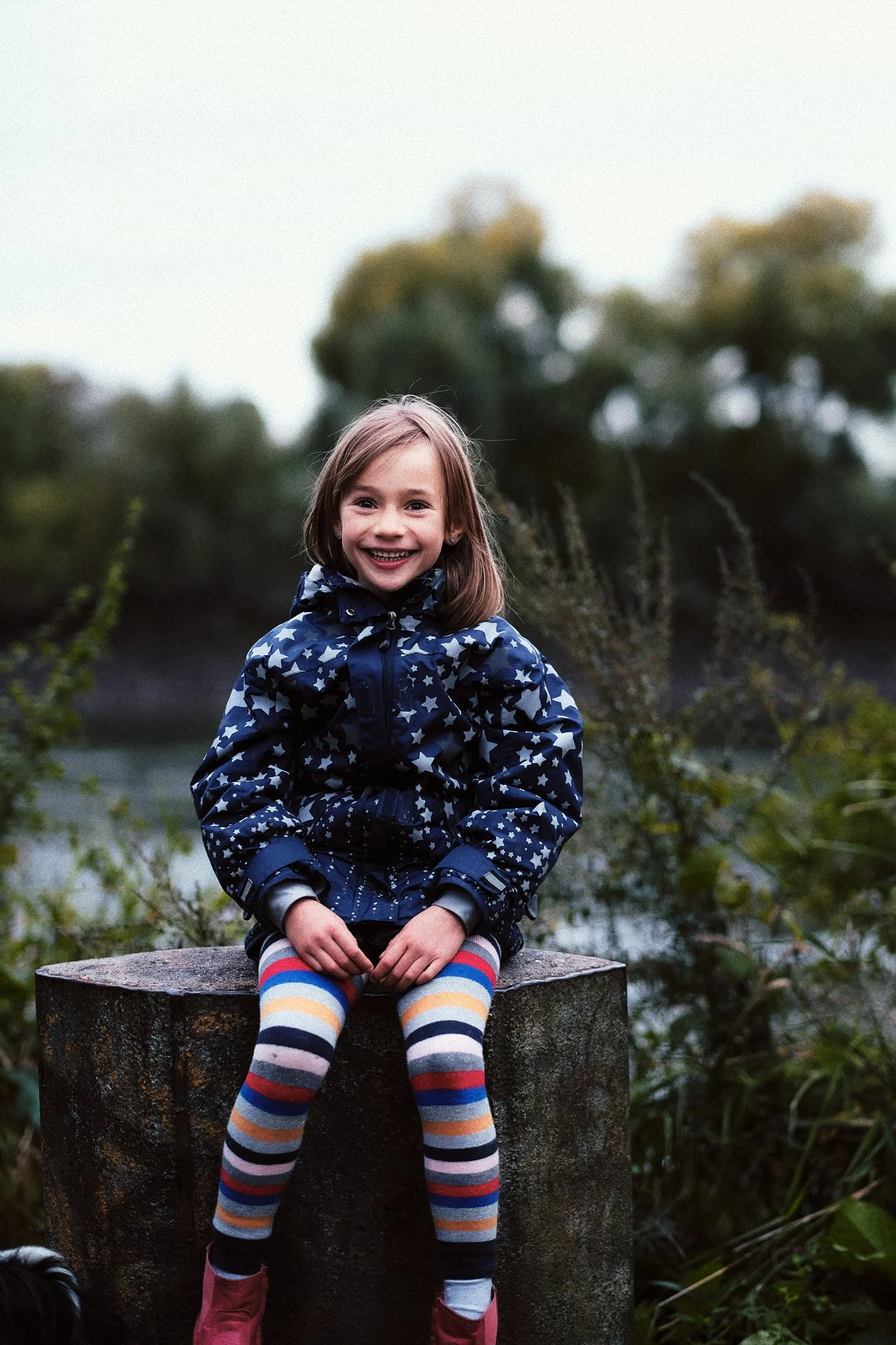 Lächelndes Mädchen mit braunem Haar, das auf einem Baumstumpf in einer natürlichen Umgebung sitzt, trägt einen blauen Mantel mit weißen Sternen und bunte gestreifte Strümpfe, im Hintergrund Bäume und Wasser