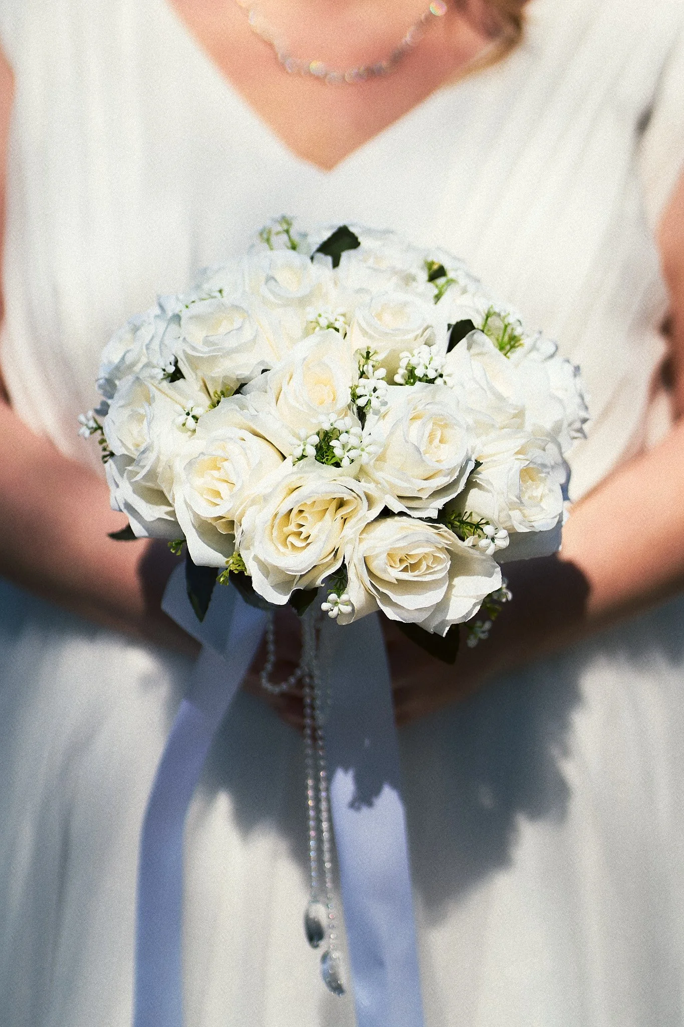 Nahaufnahme eines Brautstraußes mit weißen Rosen, kleinen weißen Blüten und grünen Blättern, dekoriert mit einer weißen Schleife und Perlen.