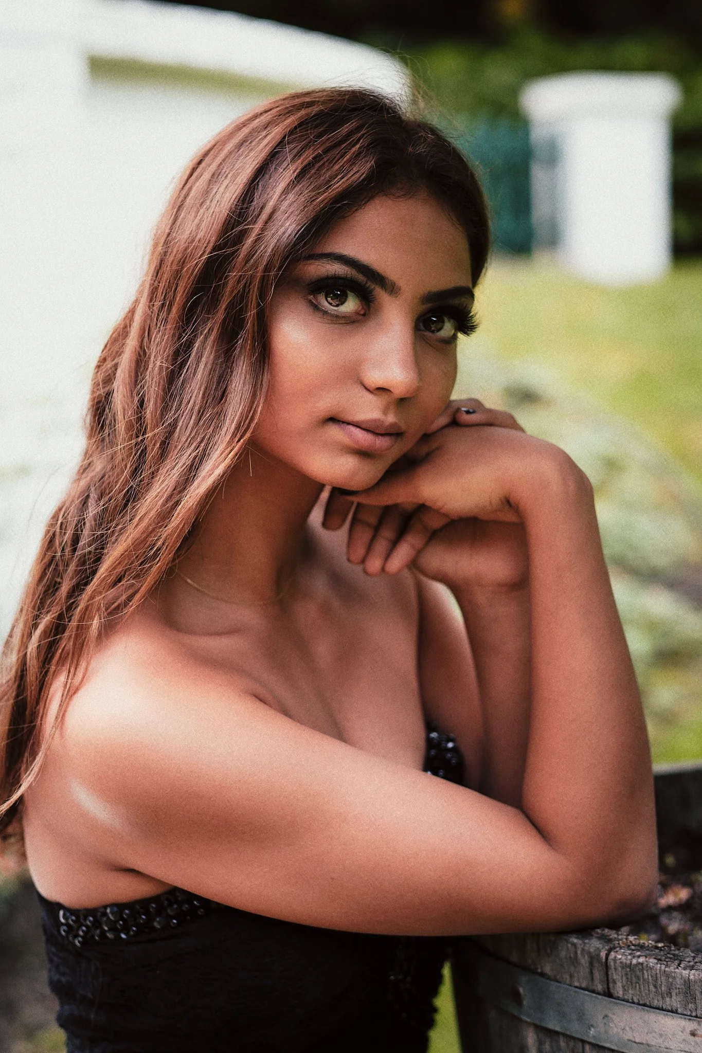 Nahaufnahme einer jungen Frau mit braunen Haaren, die auf einer Gartenbank sitzt und die Hand unter das Kinn legt, im Freien mit grünem Hintergrund.