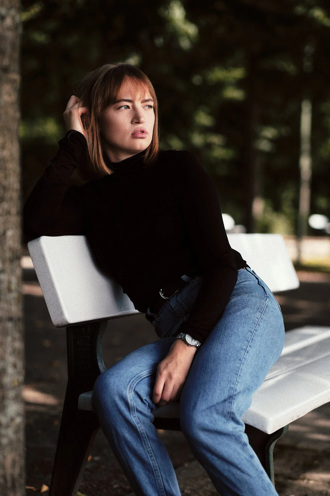 Junge Frau sitzt nachdenklich auf einer Parkbank, trägt einen schwarzen Pullover, Jeans und eine Armbanduhr, im Park mit Bäumen im Hintergrund.