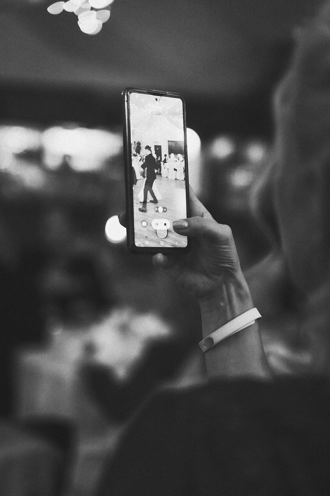 Eine Hand hält ein Smartphone, das eine Tanzveranstaltung zeigt, im Hintergrund sind bunte Lichter und mehrere Menschen zu sehen.