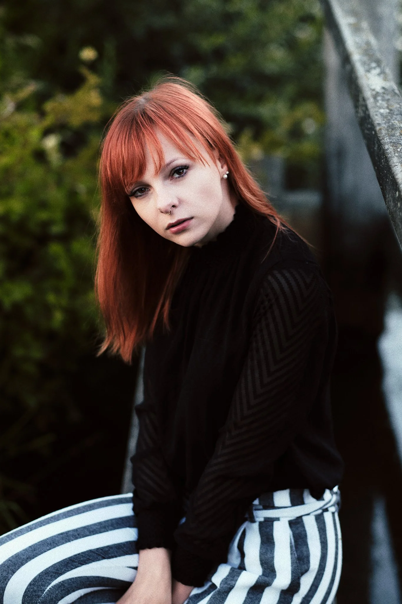 Junge Frau mit roten Haaren, dunklem Oberteil und gestreiften Hosen sitzt an einem Geländer im Freien, Blick in die Kamera gerichtet.