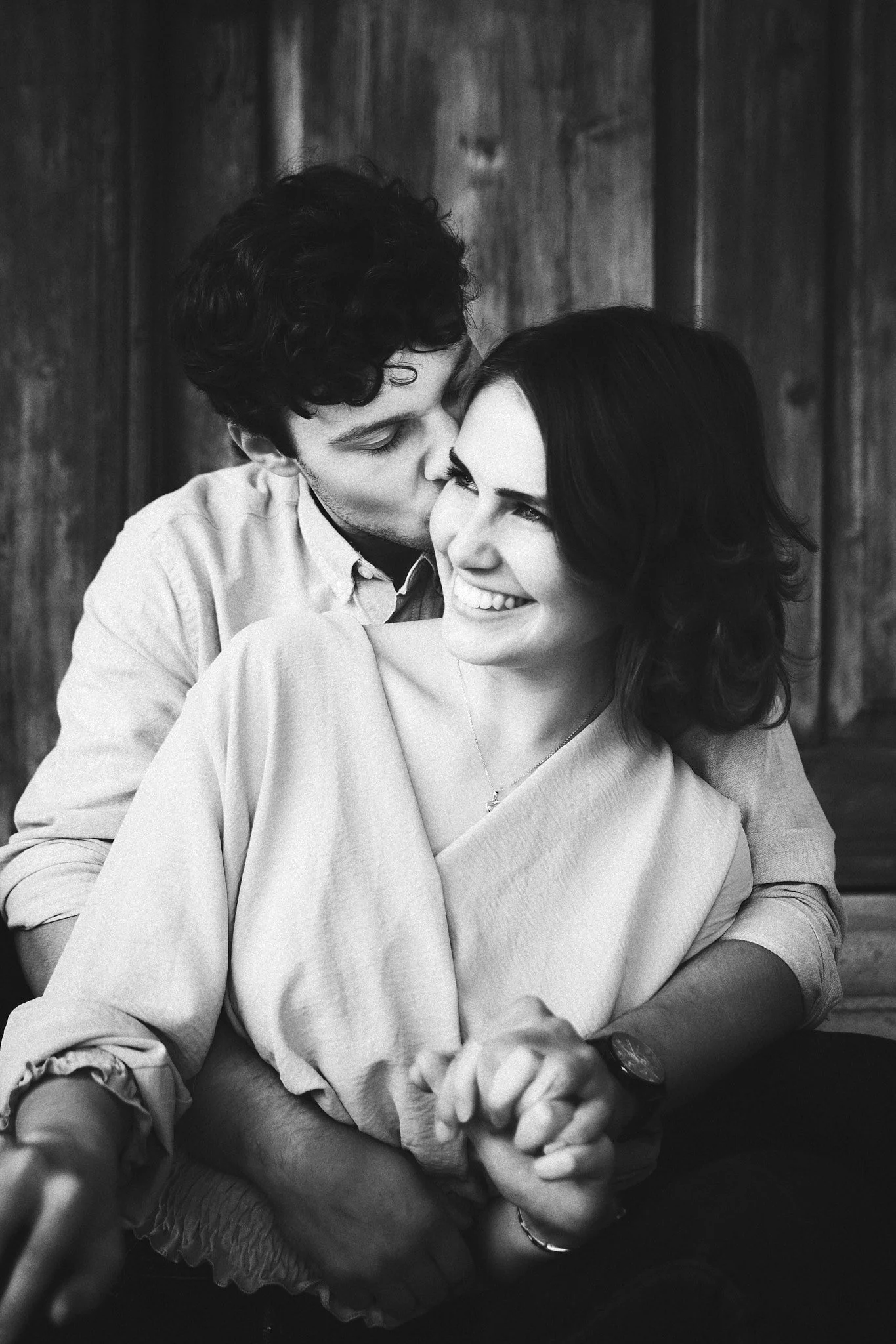 Ein lachendes Paar sitzt eng zusammen, der Mann küsst die Frau auf die Wange, im Hintergrund eine Holzvertäfelung.