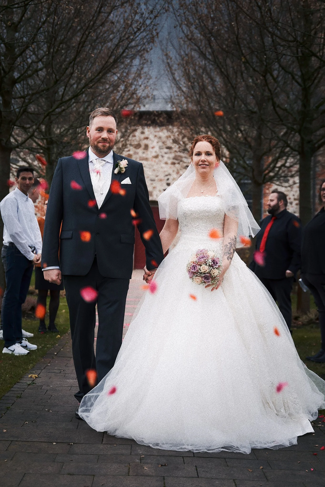 Ein Brautpaar bei einer Hochzeit im Freien, die Braut trägt ein weißes Kleid und hält einen Blumenstrauß, der Bräutigam trägt einen dunklen Anzug, im Hintergrund stehen Gäste unter Bäumen bei Sonnenuntergang, während Blütenblätter fallen.