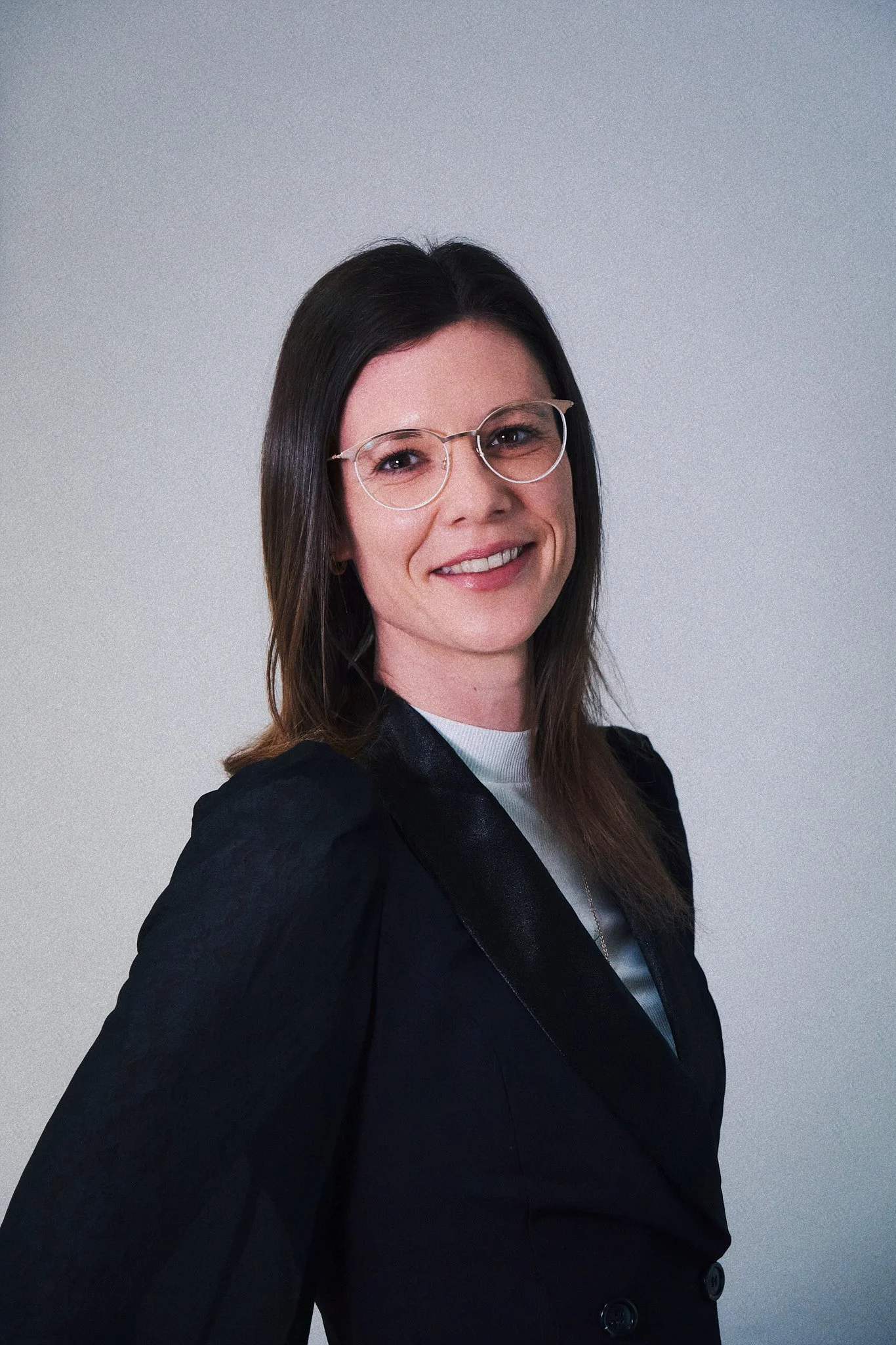 Porträt einer Frau mit schwarzen Haaren, Brille und dunklem Blazer, lächelnd, vor hellem Hintergrund.