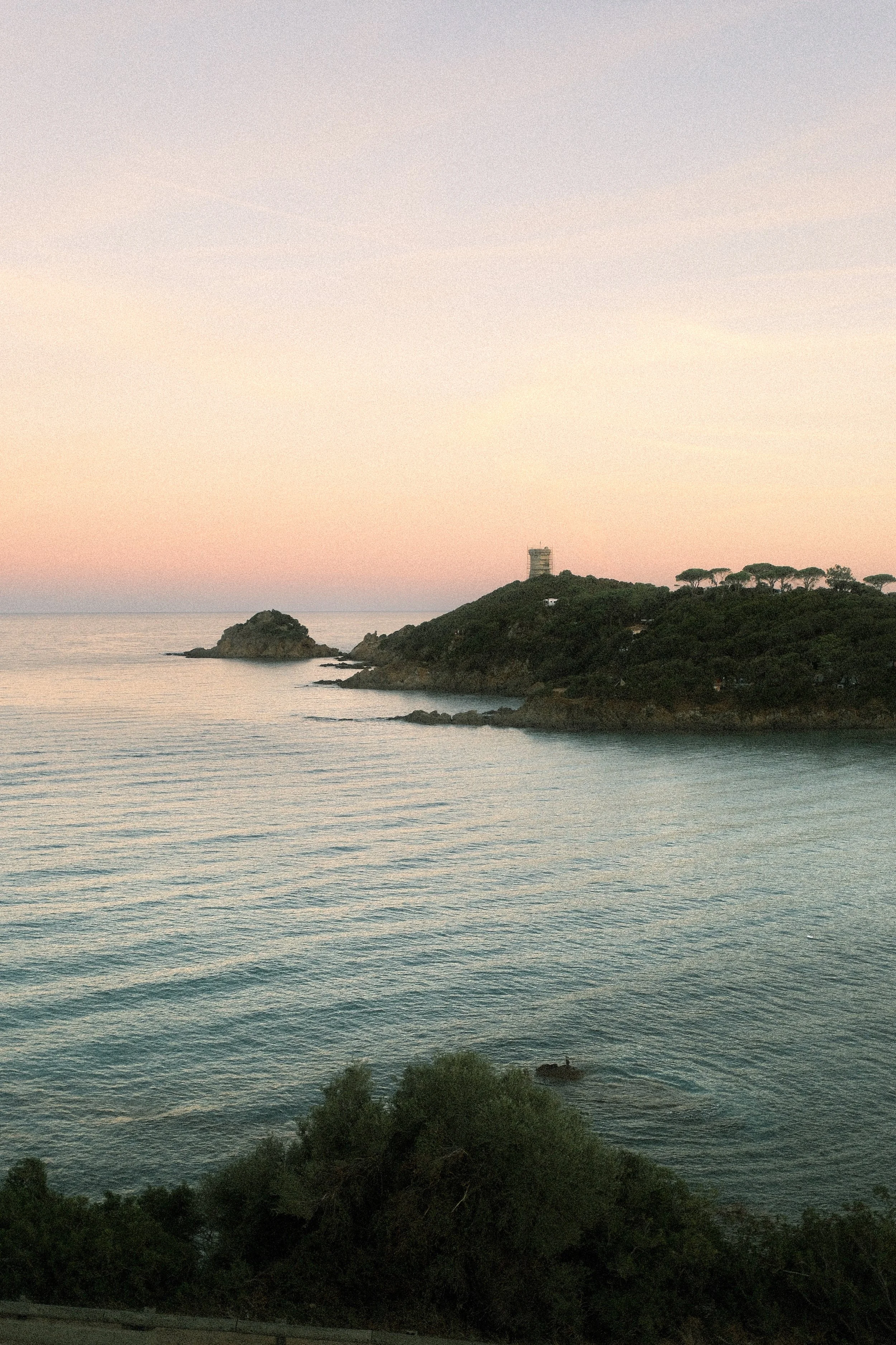 Blick auf ein ruhiges Meer bei Sonnenuntergang mit einer kleinen Insel und einem Hügel mit Bäumen und einem Gebäude auf der rechten Seite.