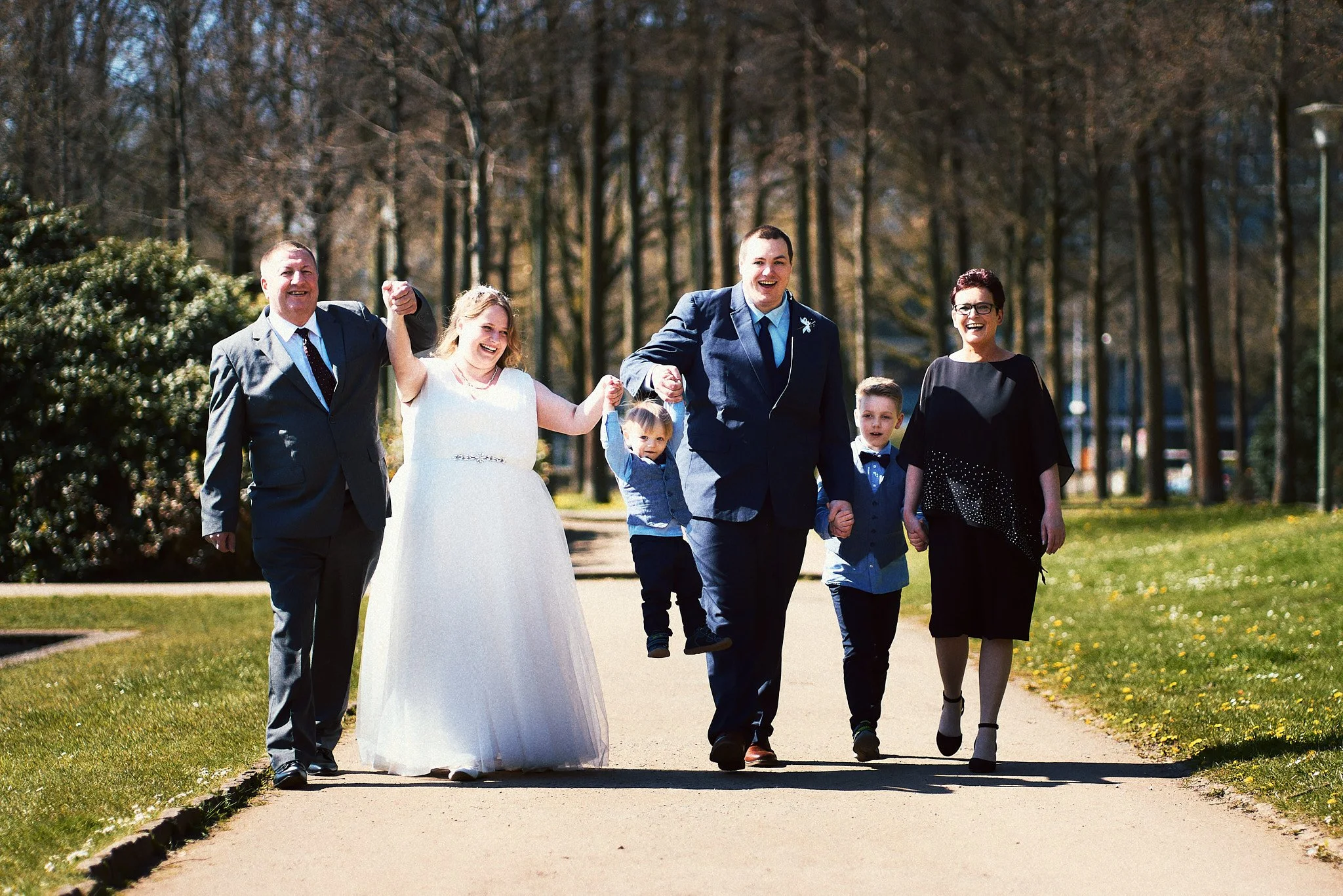 Eine Gruppe von sechs Menschen, die an einem sonnigen Tag im Park unterwegs sind. Es handelt sich um eine Hochzeitsfeier, bei der das Brautpaar mit Freunden und Familie in legerer Kleidung spazieren geht.