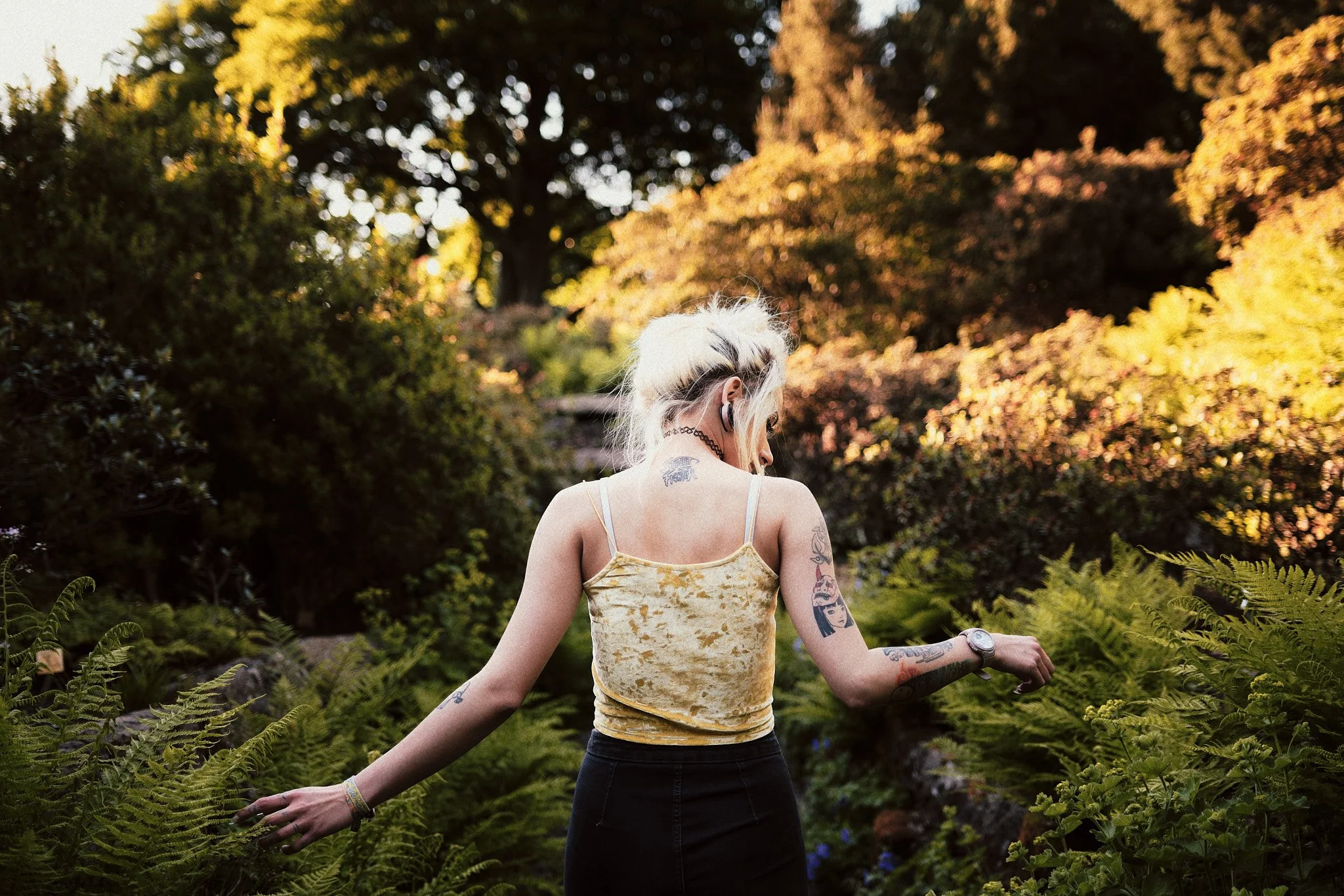 Junge Frau mit Tattoos im Garten, umgeben von grünen Pflanzen und Büschen, bei Sonnenuntergang, trägt gelben Top und schwarze Hose.