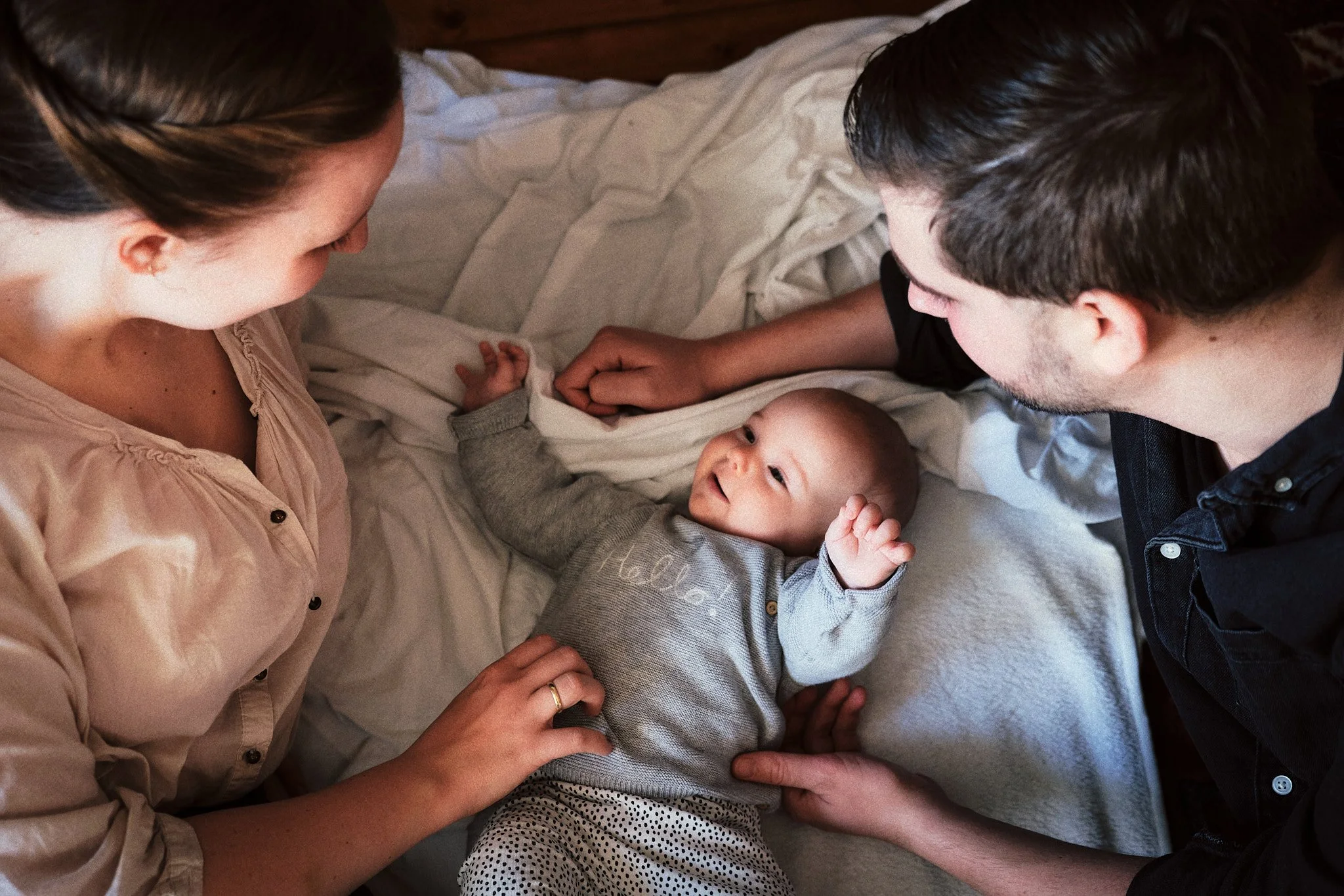 Ein Baby liegt in einem Bett, umgeben von einer Frau und einem Mann, die es liebevoll ansehen und berühren.