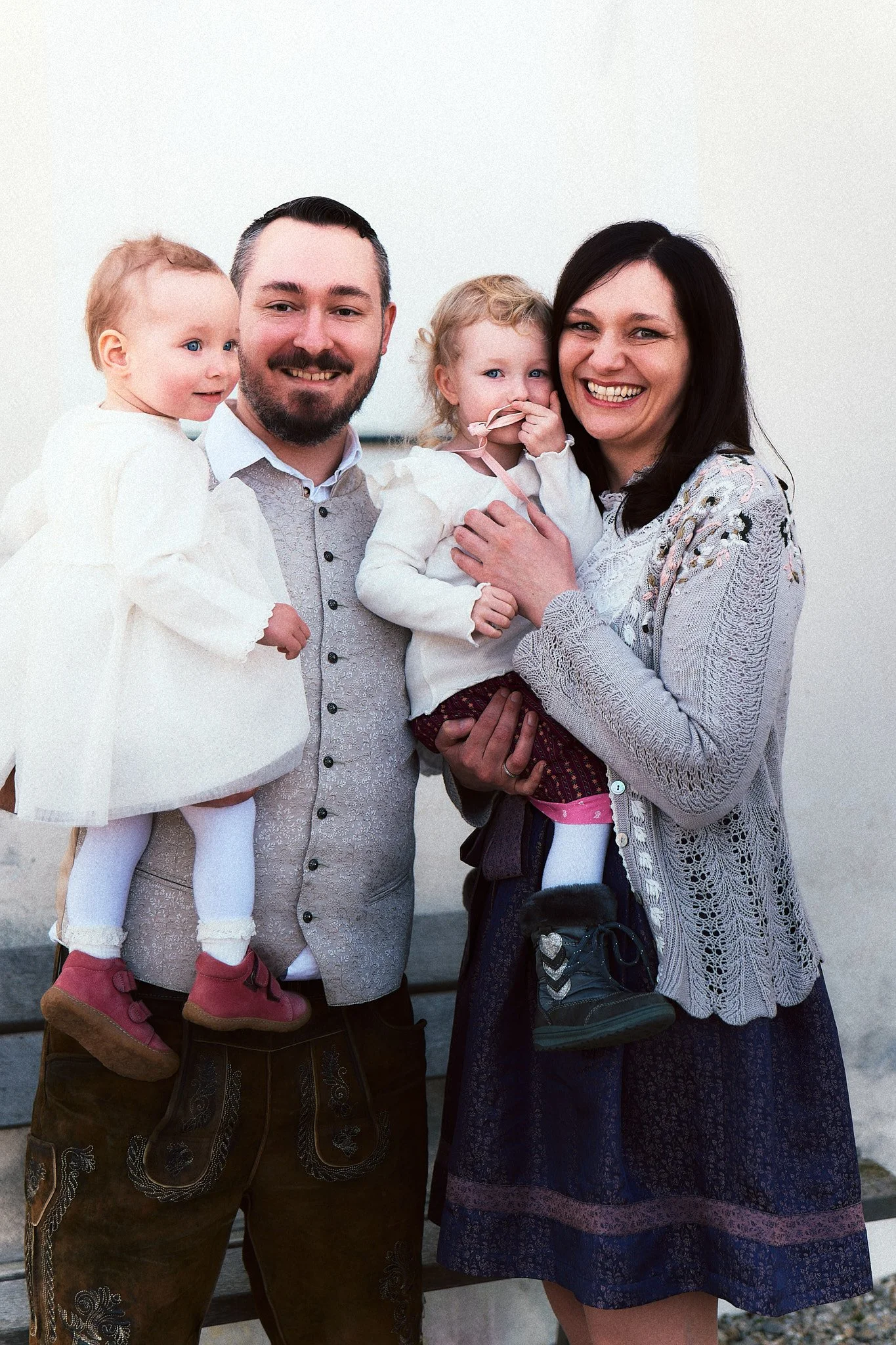 Eine glückliche Familie mit zwei Erwachsenen und zwei kleinen Mädchen, alle lächelnd, vor hellem Hintergrund.