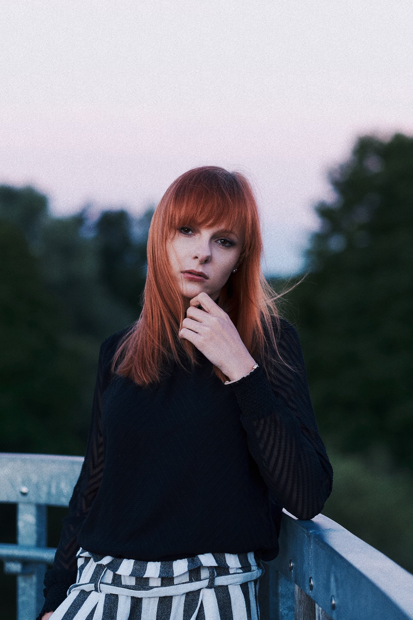 Junge Frau mit roten Haaren, dunklem Pullover, gestreiften Hosen, steht auf einer Brüstung im Freien, bei Sonnenuntergang, blickt nach vorne.