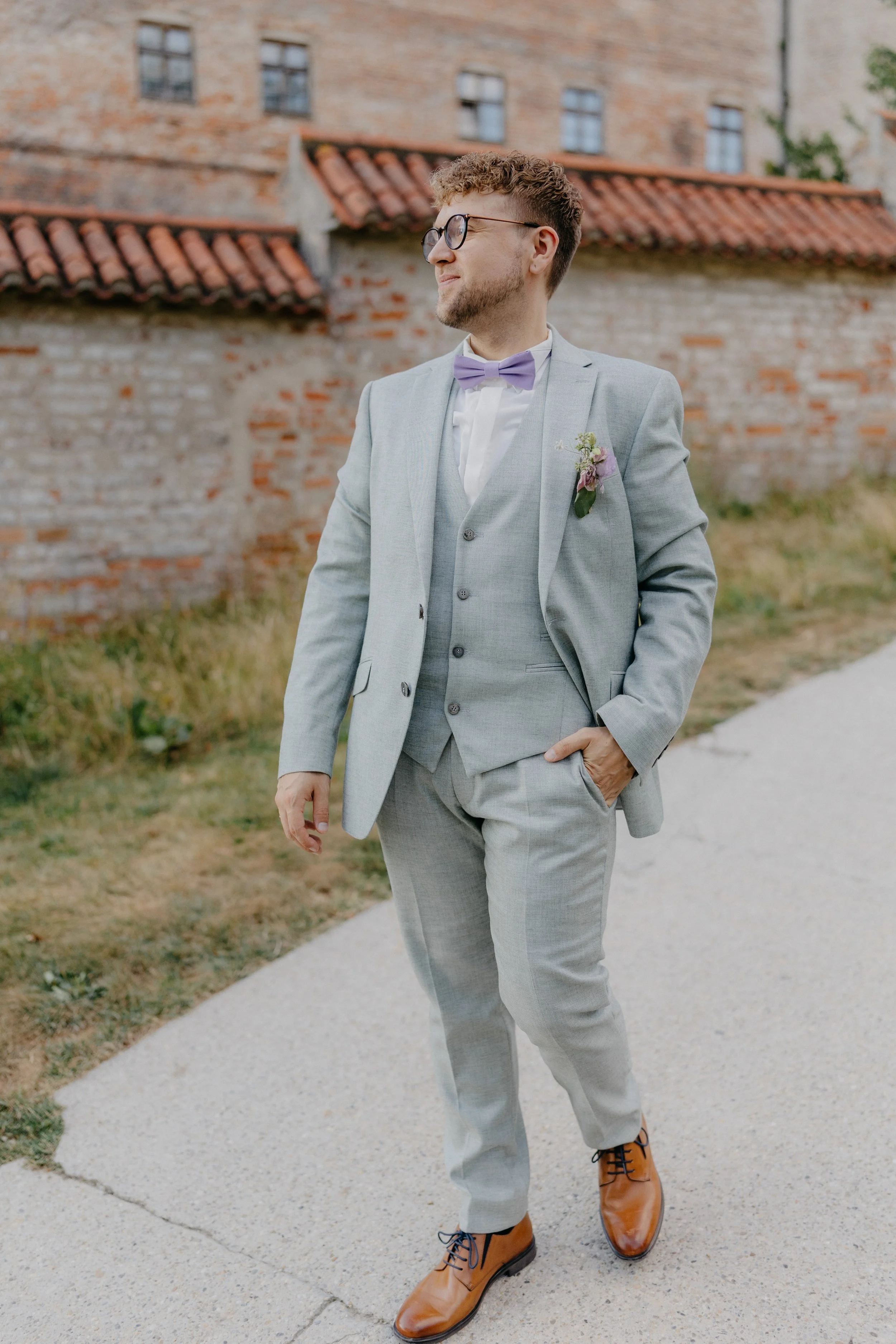 Junger Mann in grauem Anzug mit lila Fliege und Brille, trägt braune Lederschuhe, geht auf einem Weg an einer alten Backsteinmauer. Es sieht nach einer formellen Veranstaltung oder Hochzeit aus.