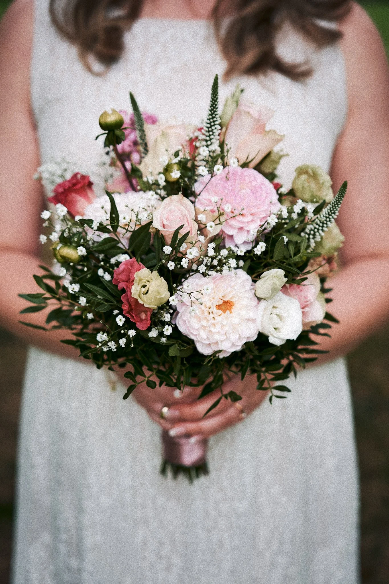 Eine Braut hält einen Blumenstrauß mit rosa, weißen und grünen Blumen vor ihrem Bauch, die im Fokus stehen, während sie ein weißes Kleid trägt.