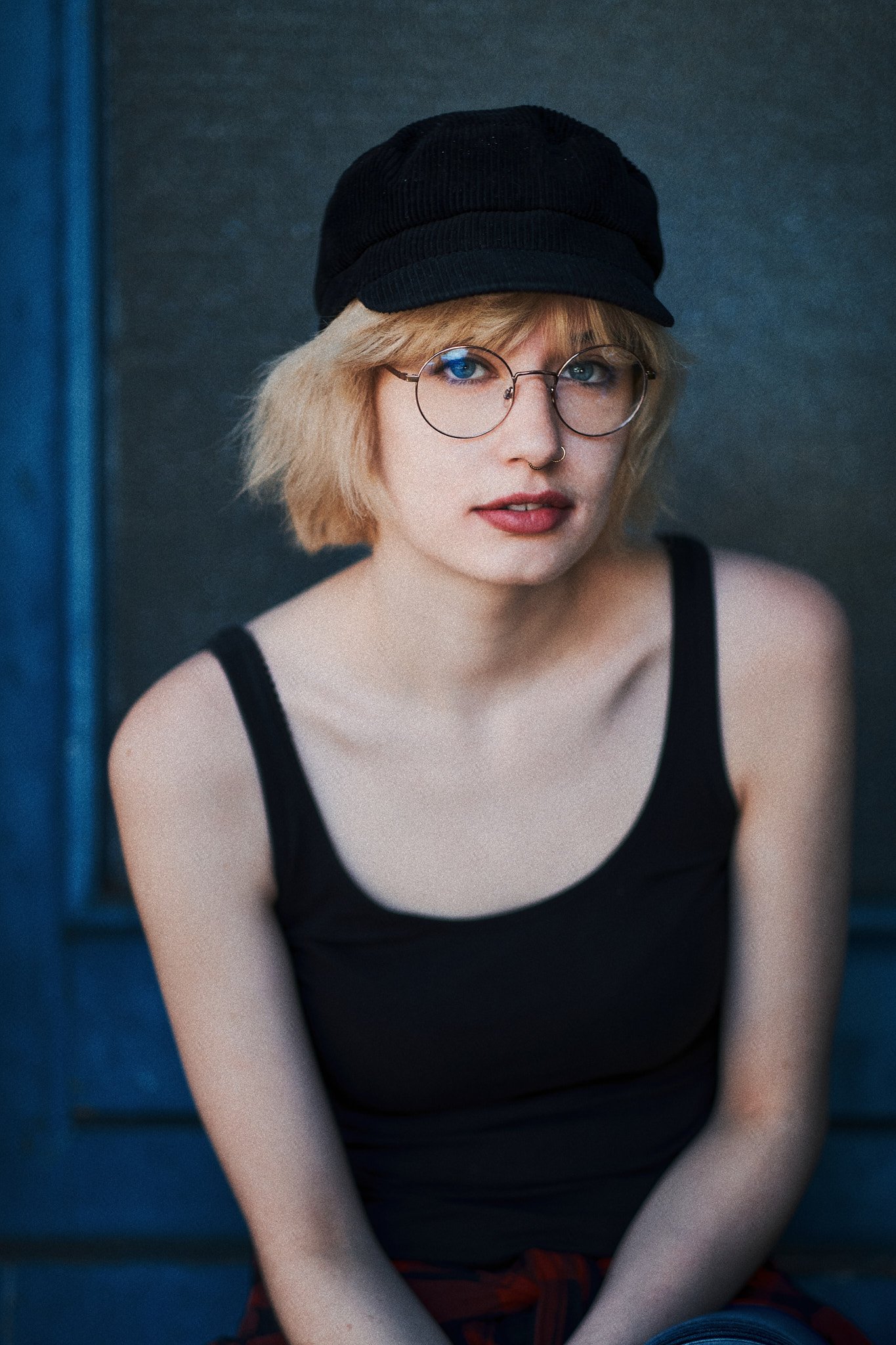 Junge Frau mit blonden, kurzen Haaren, schwarzer Kappe, runden Brillen und Nasenring sitzt gegen eine dunkle Wand in einem schwarzen Tanktop, Blick direkt in die Kamera.