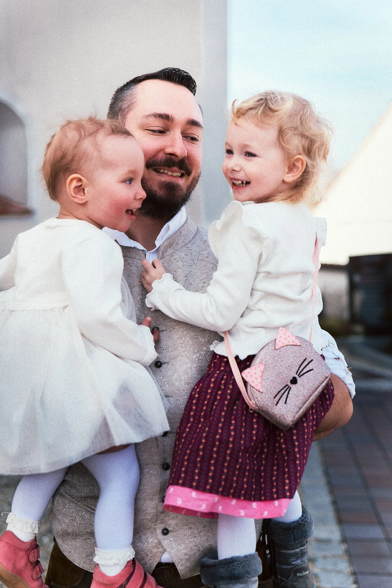 Ein Mann hält zwei kleine Mädchen in den Armen und lächelt, während sie sich umarmen. Die Mädchen tragen weiße Kleider, Strumpfhosen und Schuhe, das Mädchen auf der rechten Seite hat eine kleine Tasche mit Katzenmotiv.
