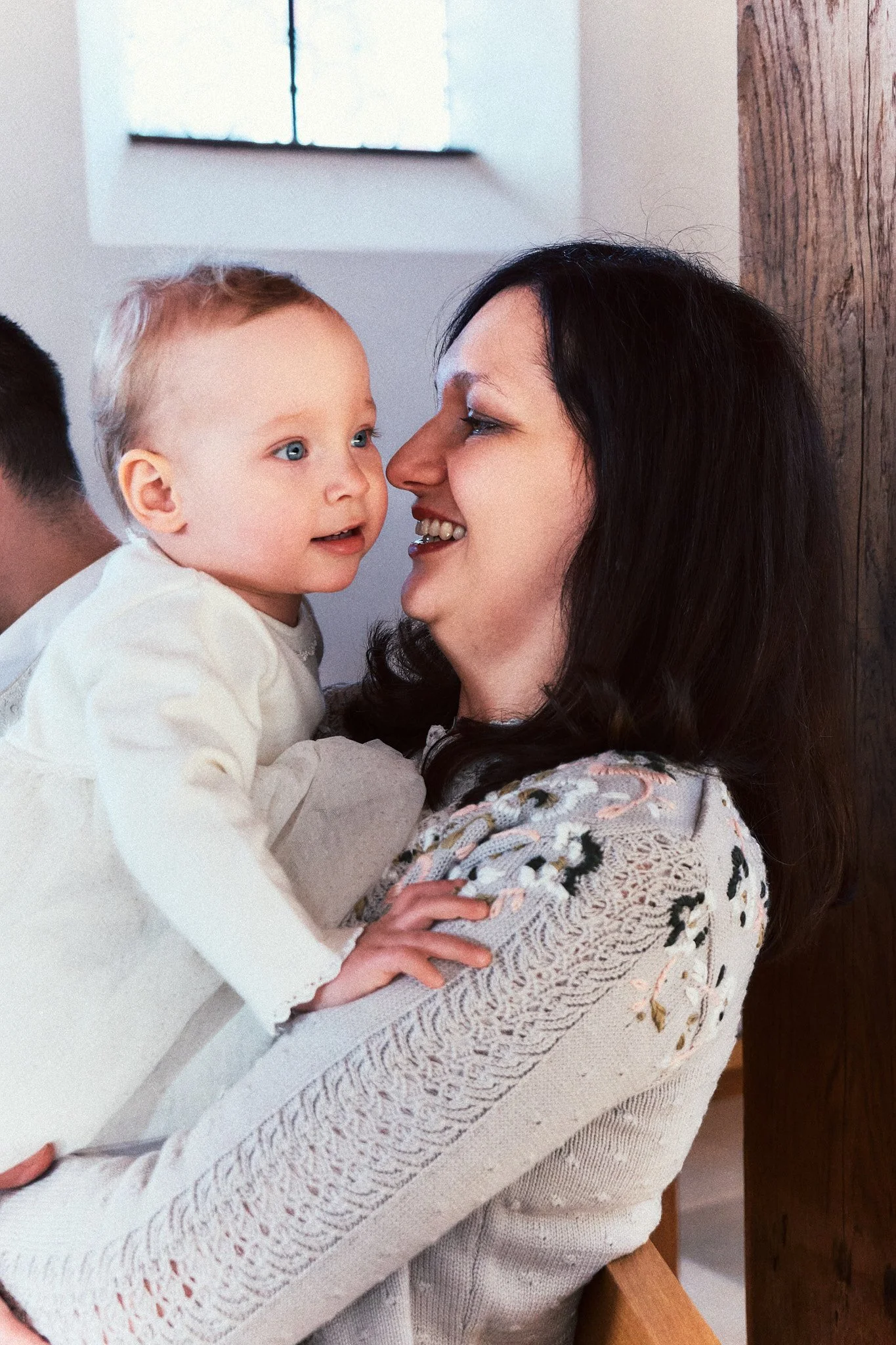 Eine Frau hält ein Baby hoch, beide schauen sich liebevoll an, im Hintergrund ein Fenster.