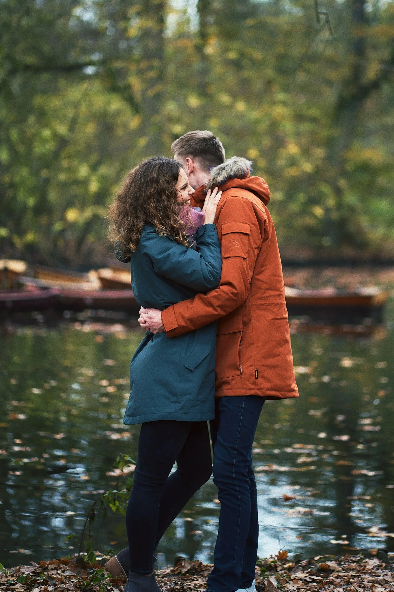 Ein Paar umarmt sich am Wasser in einem Wald, Herbstlaub am Boden, bunte Blätter an den Bäumen.
