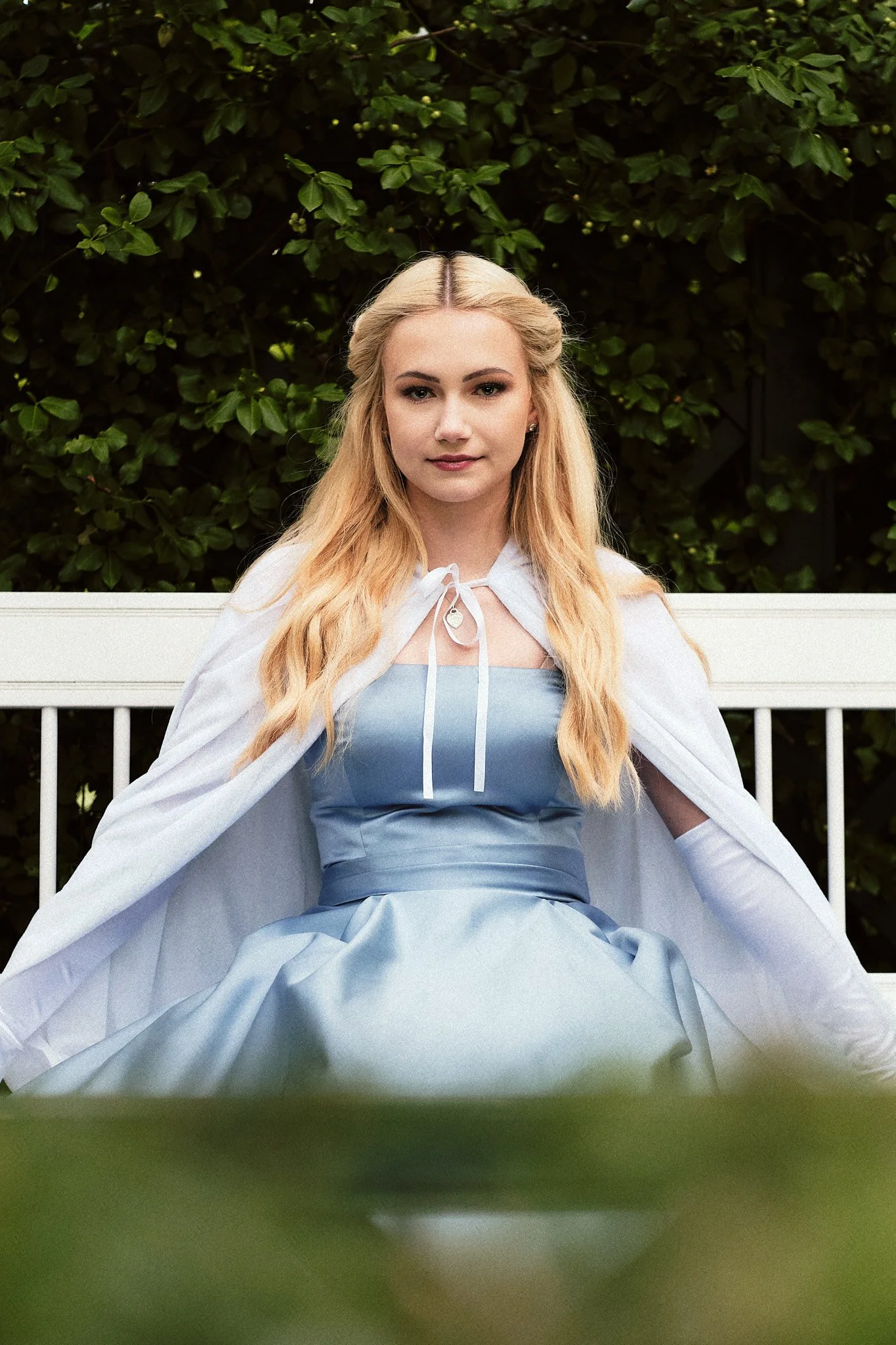 Junge Frau in blauer Kleider und weißer Umhängetunika, sitzend auf einer weißen Gartenbank, vor einer grünen Hecke.