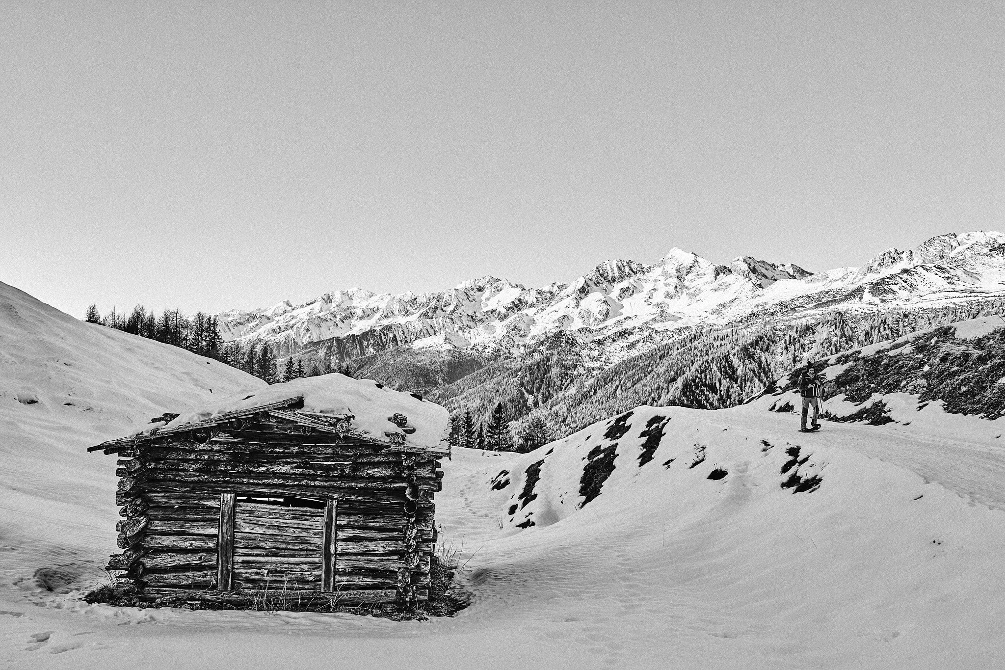 Ein einsames, hölzernes Blockhaus in verschneiter Berglandschaft mit schneebedeckten Bergen im Hintergrund, eine Person mit Skiern auf dem Weg im Schnee.