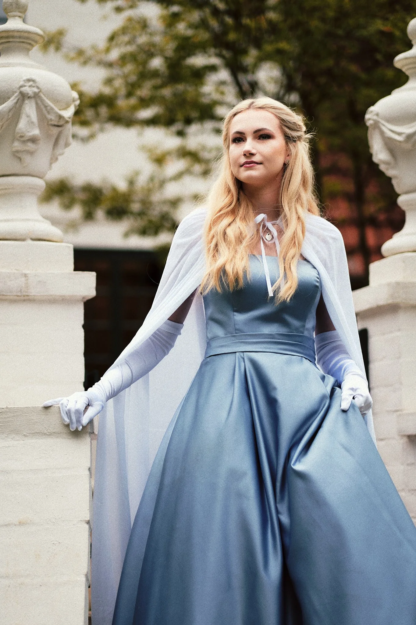 Junge Frau in einem eleganten blauen Kleid und weißen Handschuhen steht vor einer weißen Metallbrüstung, umgeben von Bäumen mit Herbstlaub.