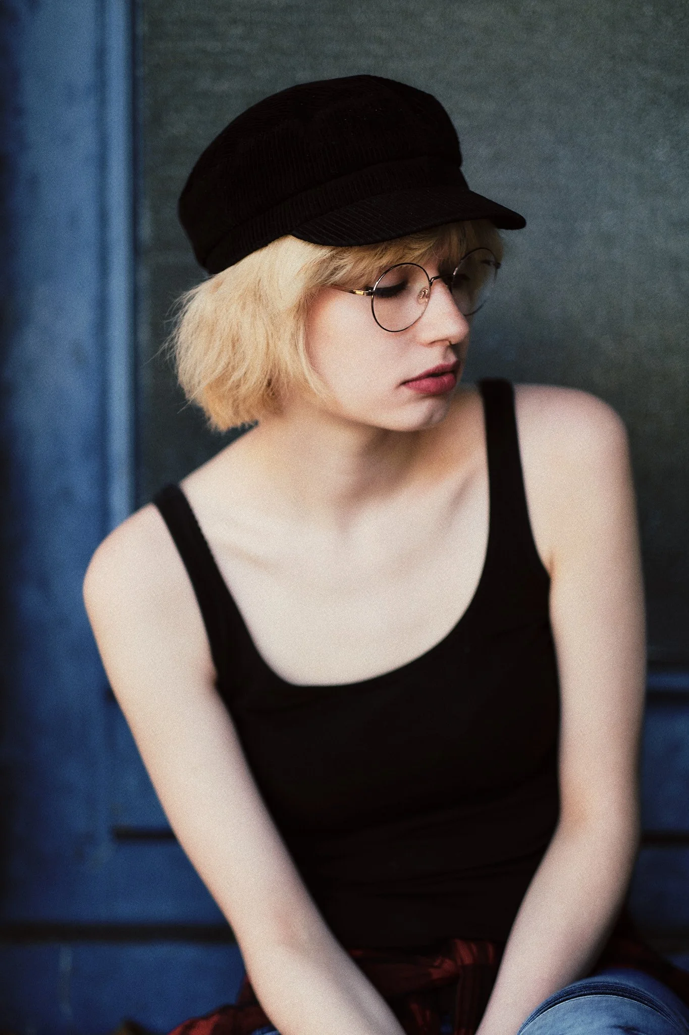 Junge Frau mit rotem Lippenstift, Brille, schwarzem Hut und schwarzem Top sitzt nachdenklich gegen eine blaue Wand.