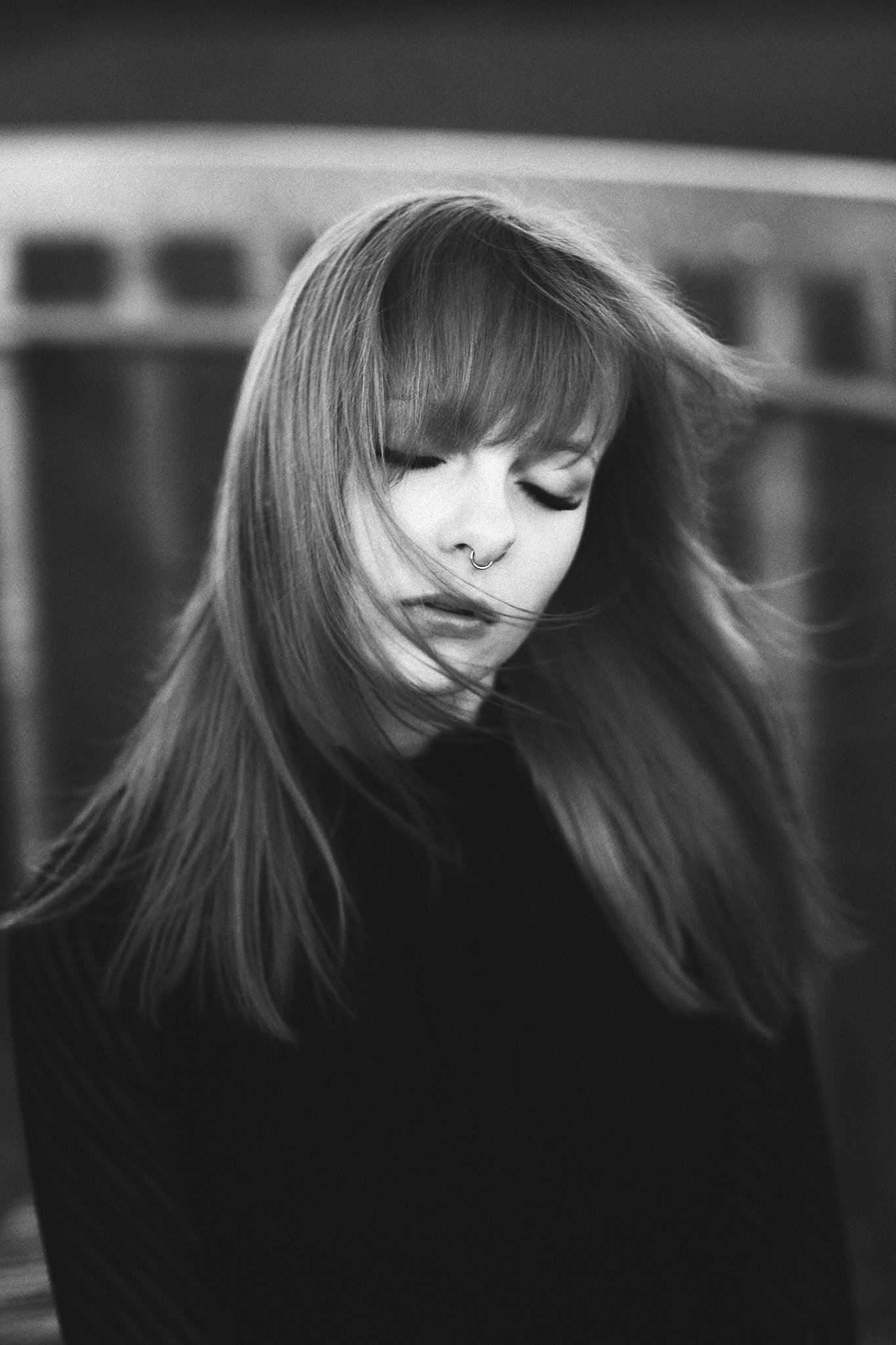 Schwarz-weiß Foto einer jungen Frau mit langen Haaren, vom Wind zerzaust, mit Nasenpiercing, geschlossenen Augen und einem nachdenklichen Ausdruck.