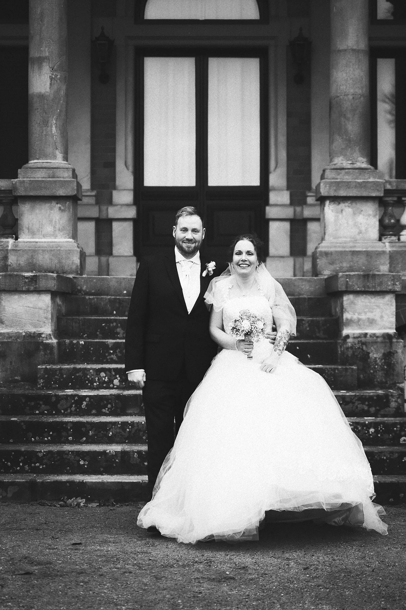 Ein Hochzeitsfoto eines Paares in formeller Kleidung, das vor einem historischen Gebäude auf den Stufen steht, schwarz-weiß aufgenommen.