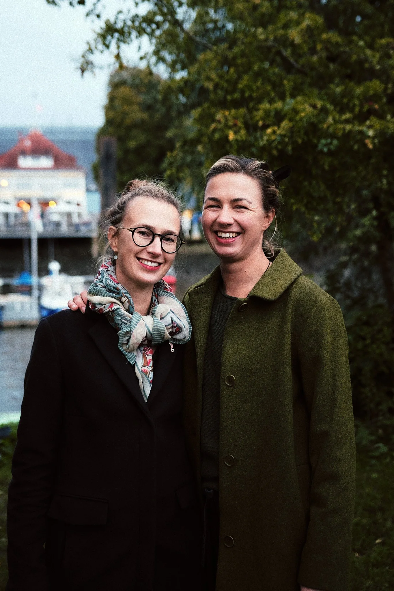 Zwei lachende Frauen stehen nebeneinander draußen, mit Bäumen und einem Wasserweg im Hintergrund.