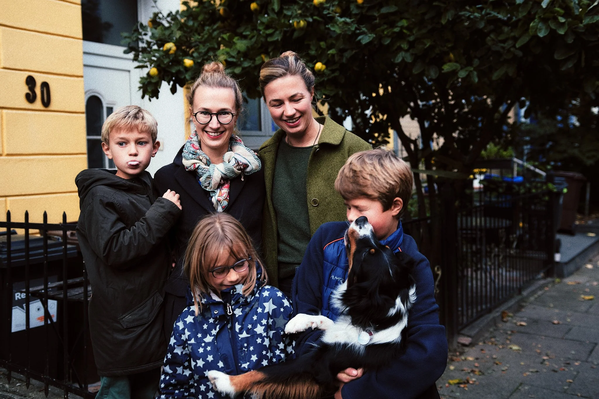 Familie mit drei Kindern und einem Hund draußen auf der Straße, im Hintergrund ein Baum und Häuser.