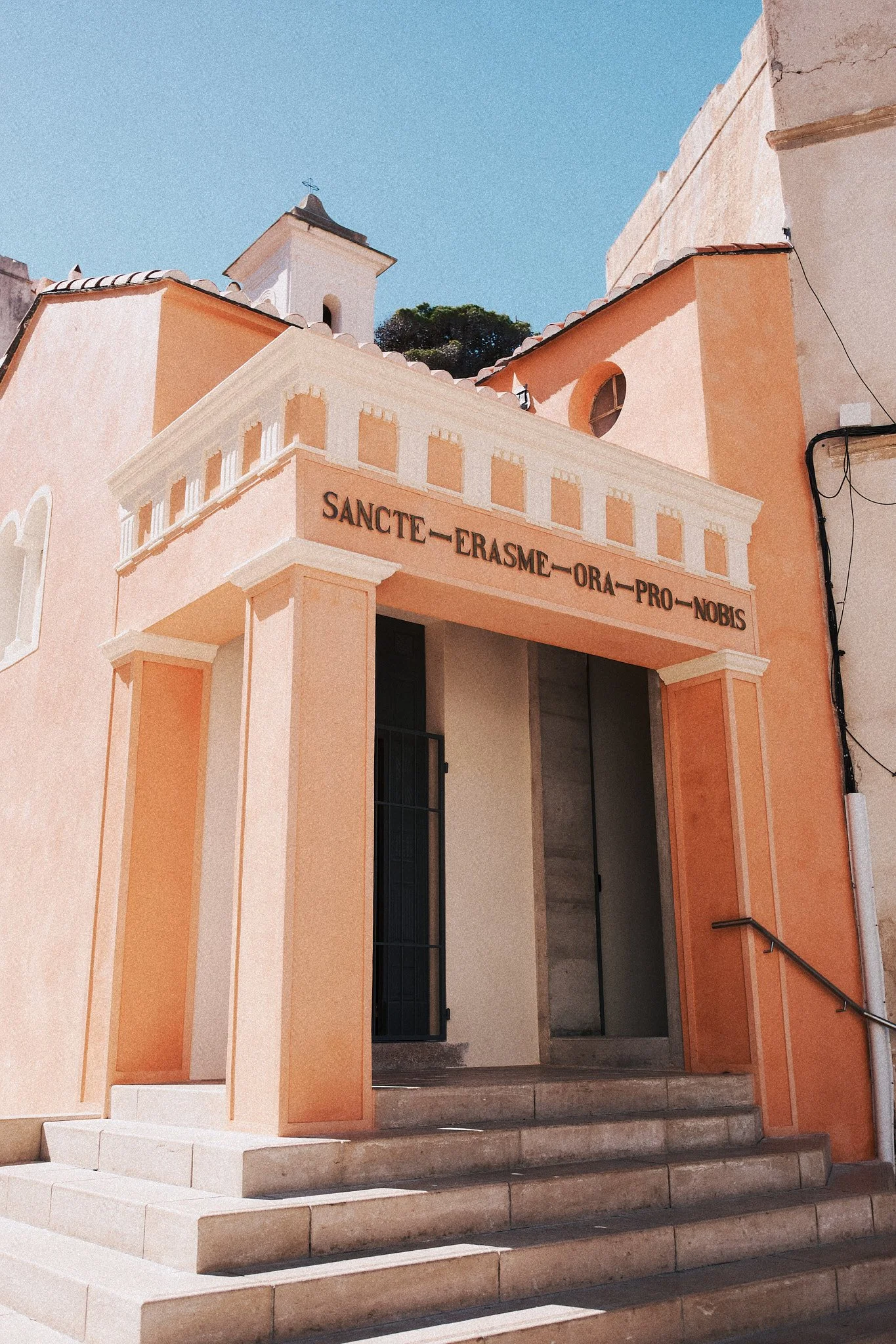 Ein hell rosa bis orangefarbener Kircheneingang mit Stufen und einem Säulenportal. Über dem Eingang steht ein lateinischer Spruch: "SANCTE ERASME ORA PRO NOBIS." Im Hintergrund sind Dächer und ein blauer Himmel sichtbar.