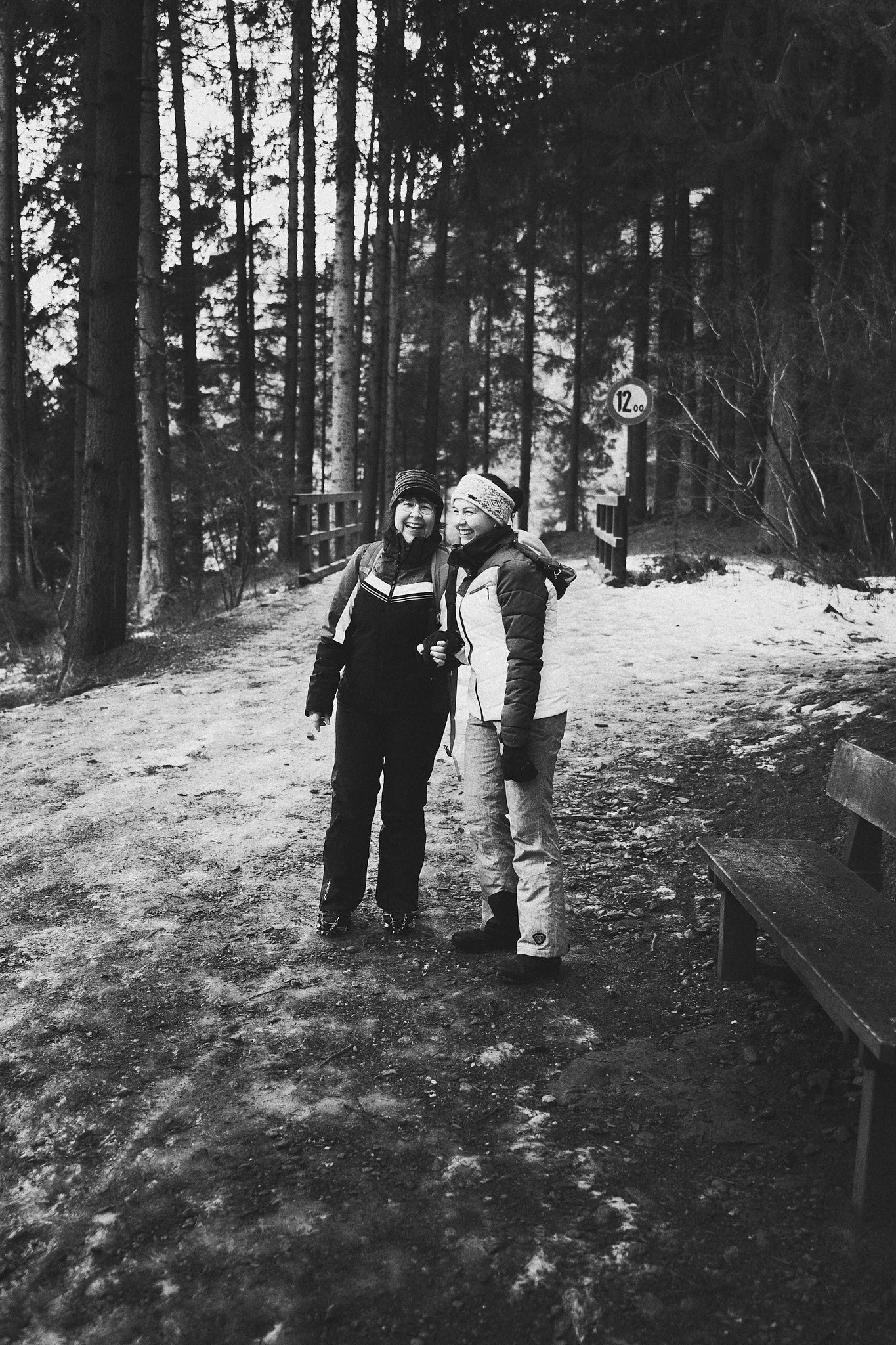 Zwei lachende Frauen in Winterkleidung stehen auf einem verschneiten Weg in einem Wald, im Hintergrund Bäume und eine Geschwindigkeitsbegrenzung '12.00'.