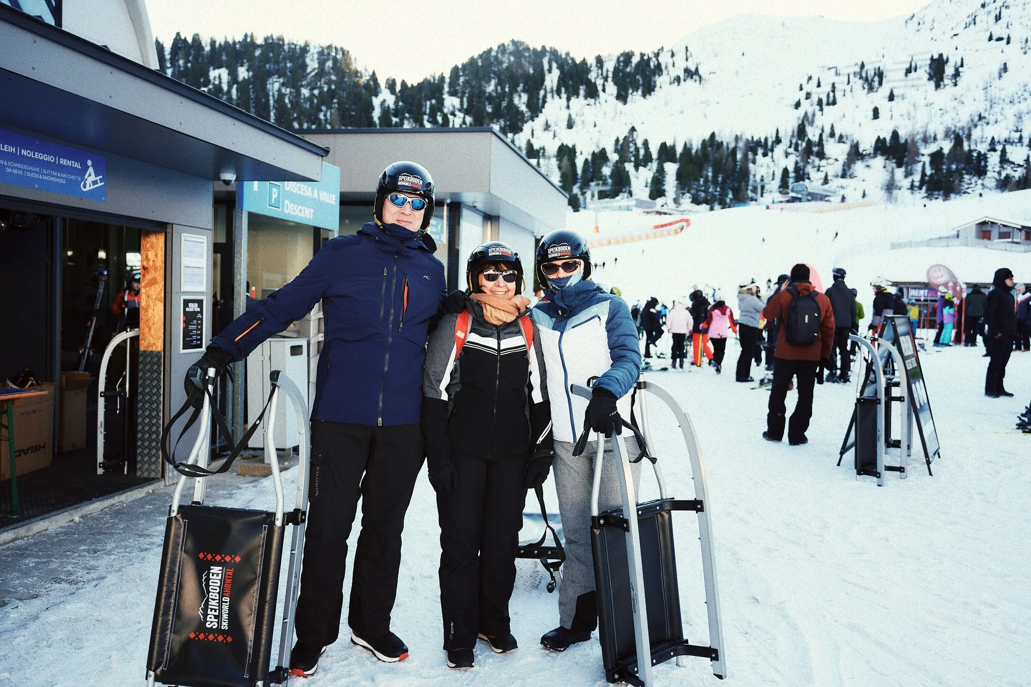 Drei Skifahrer vor einem Skilift in einer verschneiten Bergregion mit anderen Skifahrern und Skigebäuden im Hintergrund.