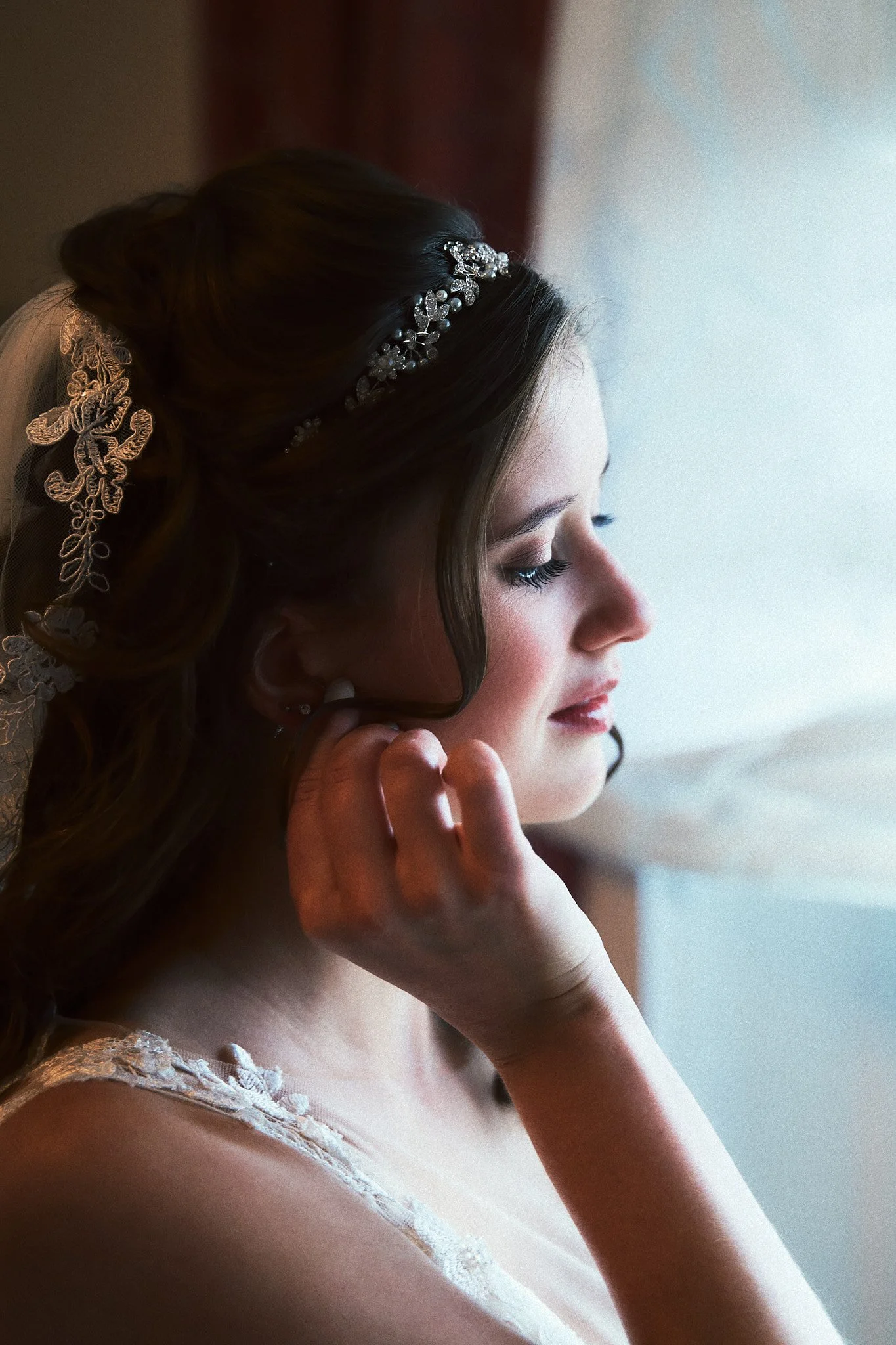 Braut mit eleganter Hochsteckfrisur und Perlen-Haaraccessoire, die ihre Ohrringe anpasst, blickt nach draußen durch ein Fenster.