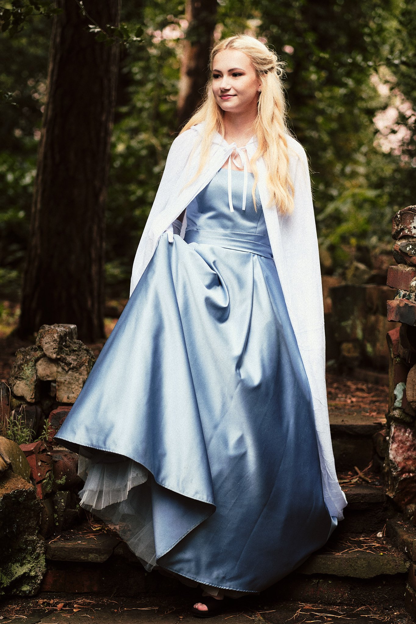 Eine junge Frau trägt ein langes, hellblaues Kleid, einen weißen Umhang und steht im Grünen.