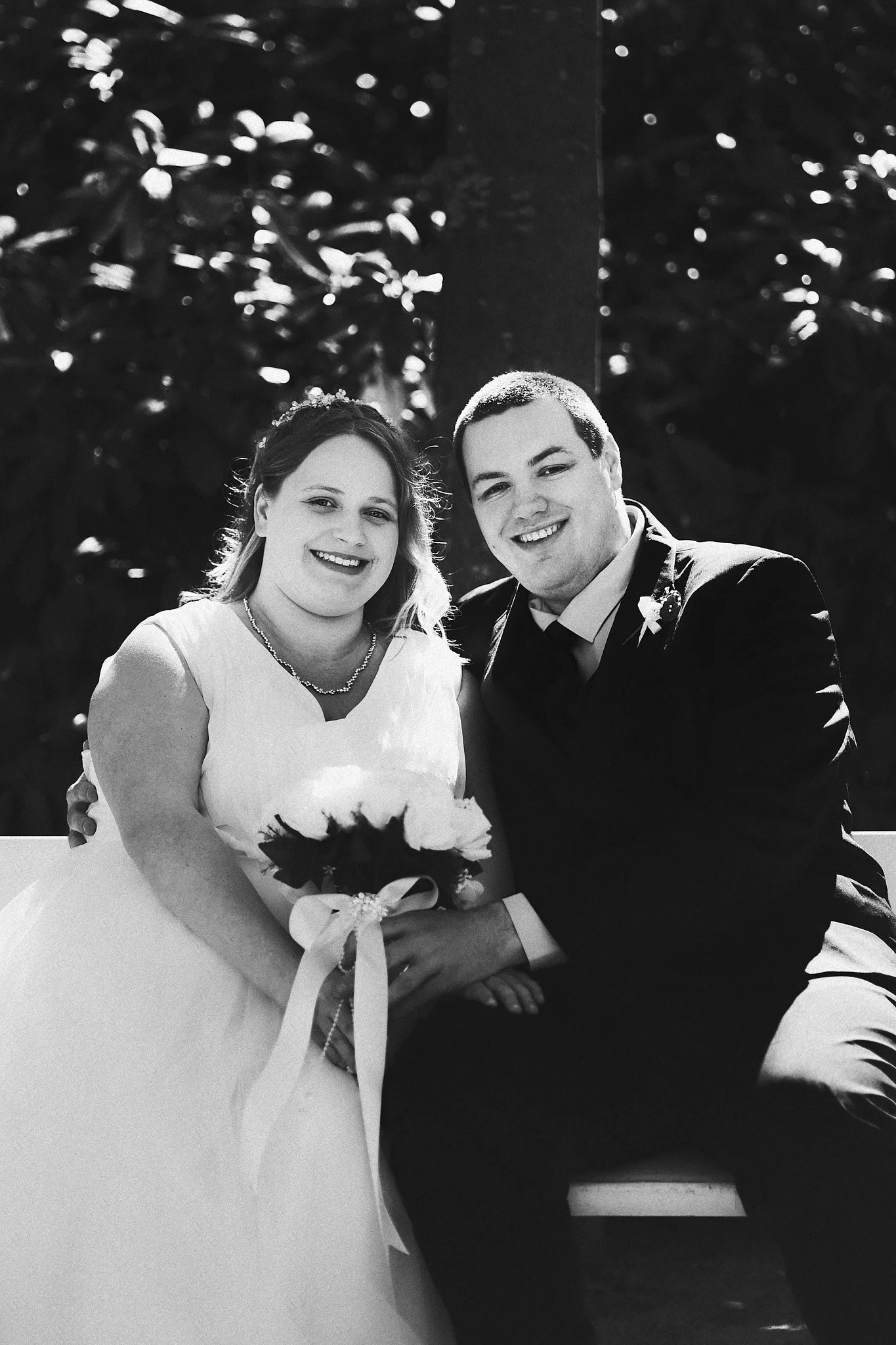 Braut und Bräutigam bei ihrer Hochzeit, sitzend und lächelnd, im Schwarz-Weiß-Foto. Sie halten einen Blumenstrauß. Hintergrund mit Blättern und Bäumen.